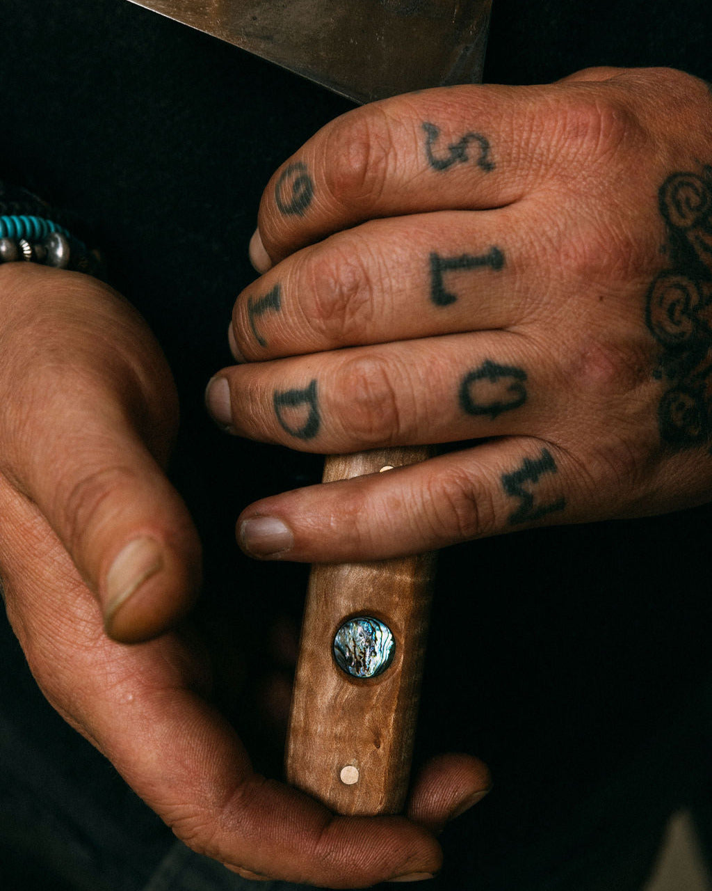 Close-up of tattooed hands holding a wooden cleaver handle with an abalone inlay on the handle