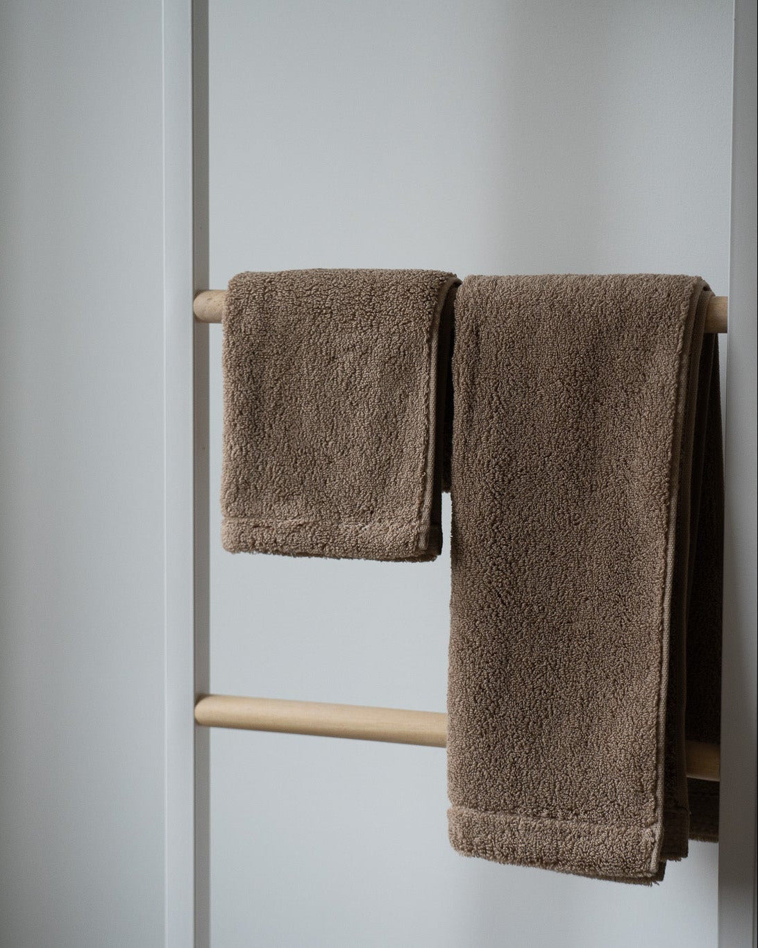 Two brown towels hanging on a wooden towel rack against a light gray wall.