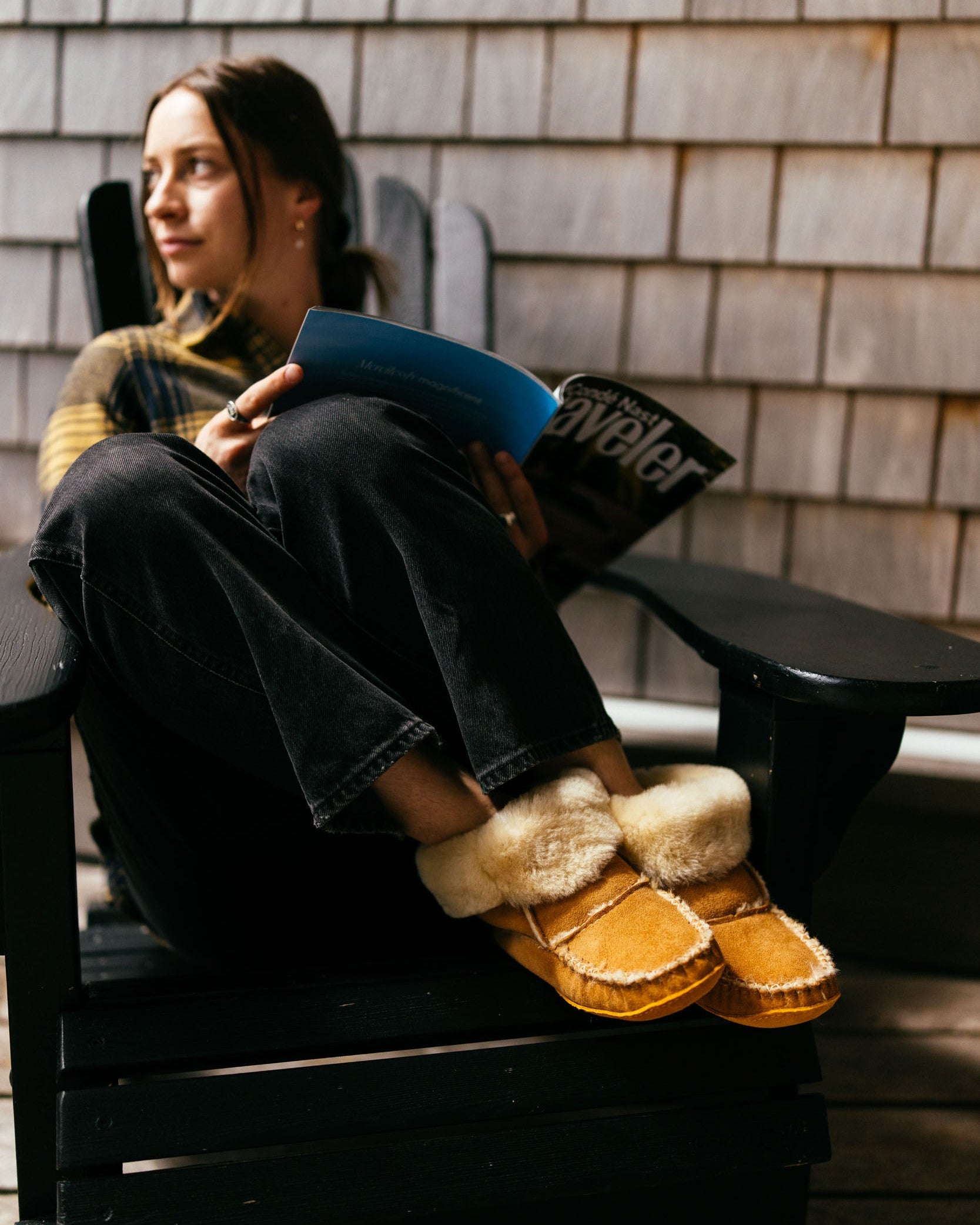 Person sitting on a chair with a book and a magazine, wearing slippers, against a wooden wall.