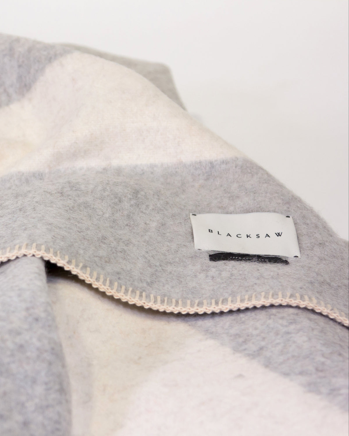 A close-up of a soft woven blanket with a light gray and off-white check pattern, featuring cream-colored stitched edges and a small white brand label that reads 'BLACKAW'.