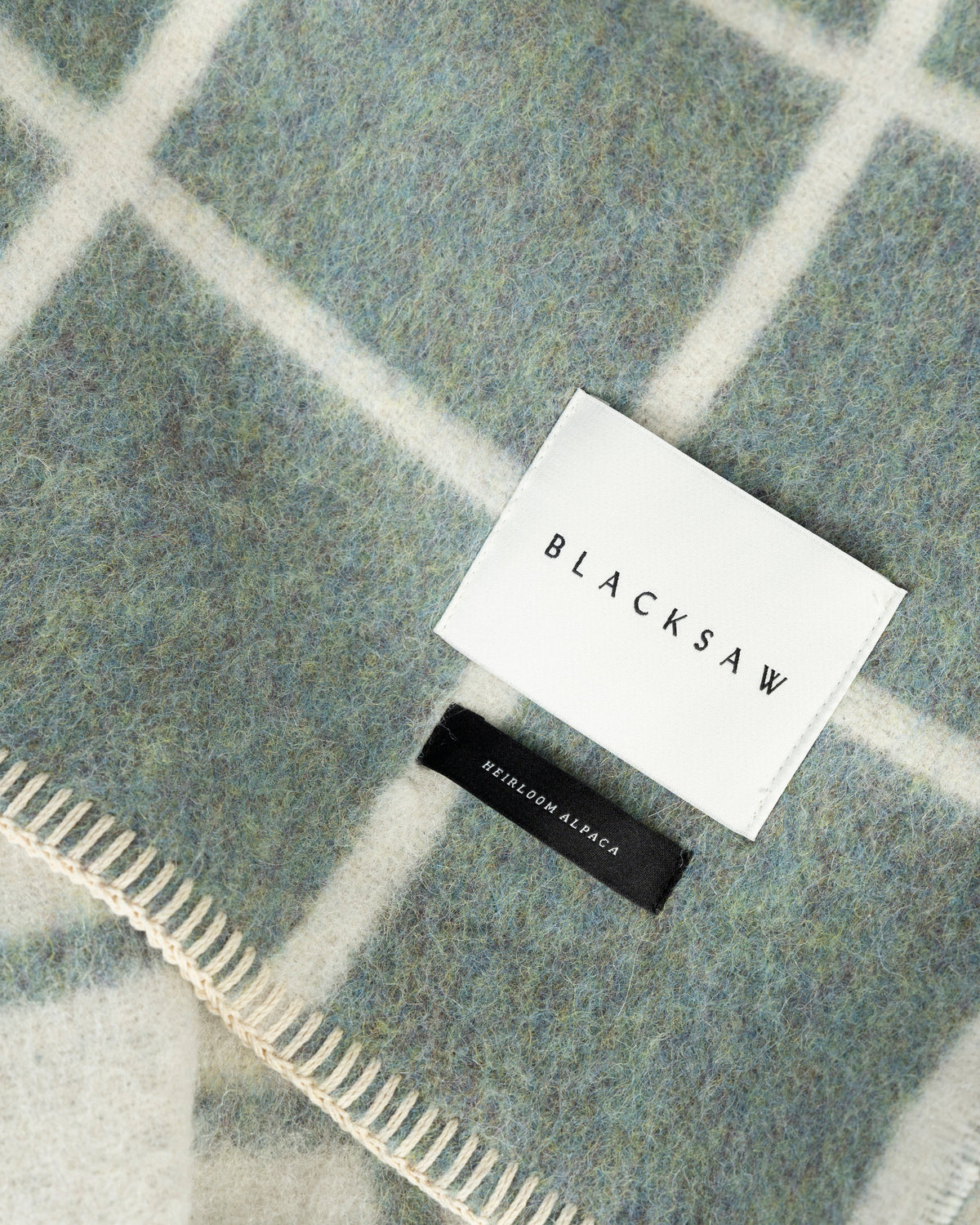 A close-up of a soft woven throw blanket with a muted blue-gray and white windowpane plaid pattern, featuring a white sewn label that reads BLACKSAW and a small black brand tag.