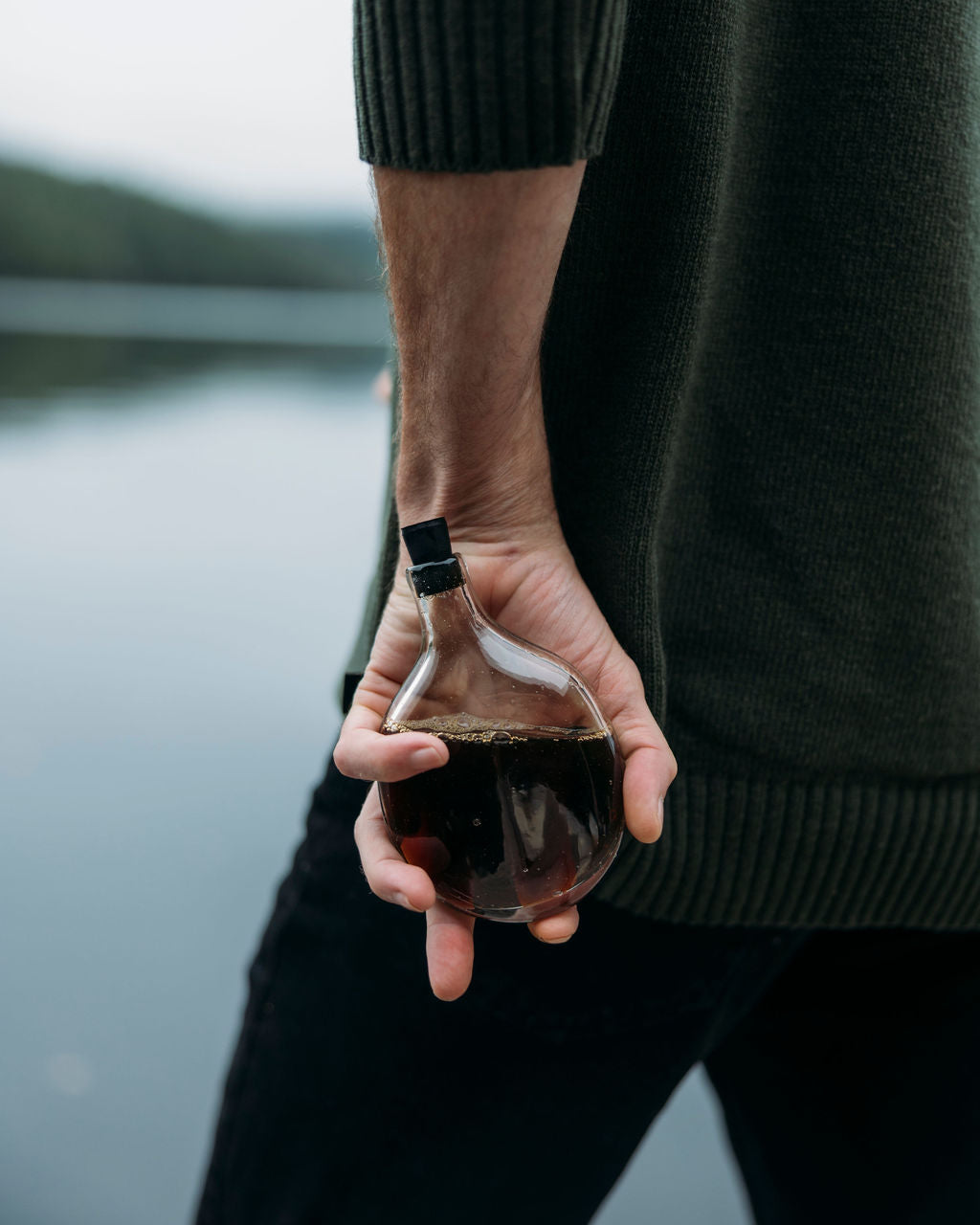 Person holding a Goodbeast glass flask with black cap against a scenic background