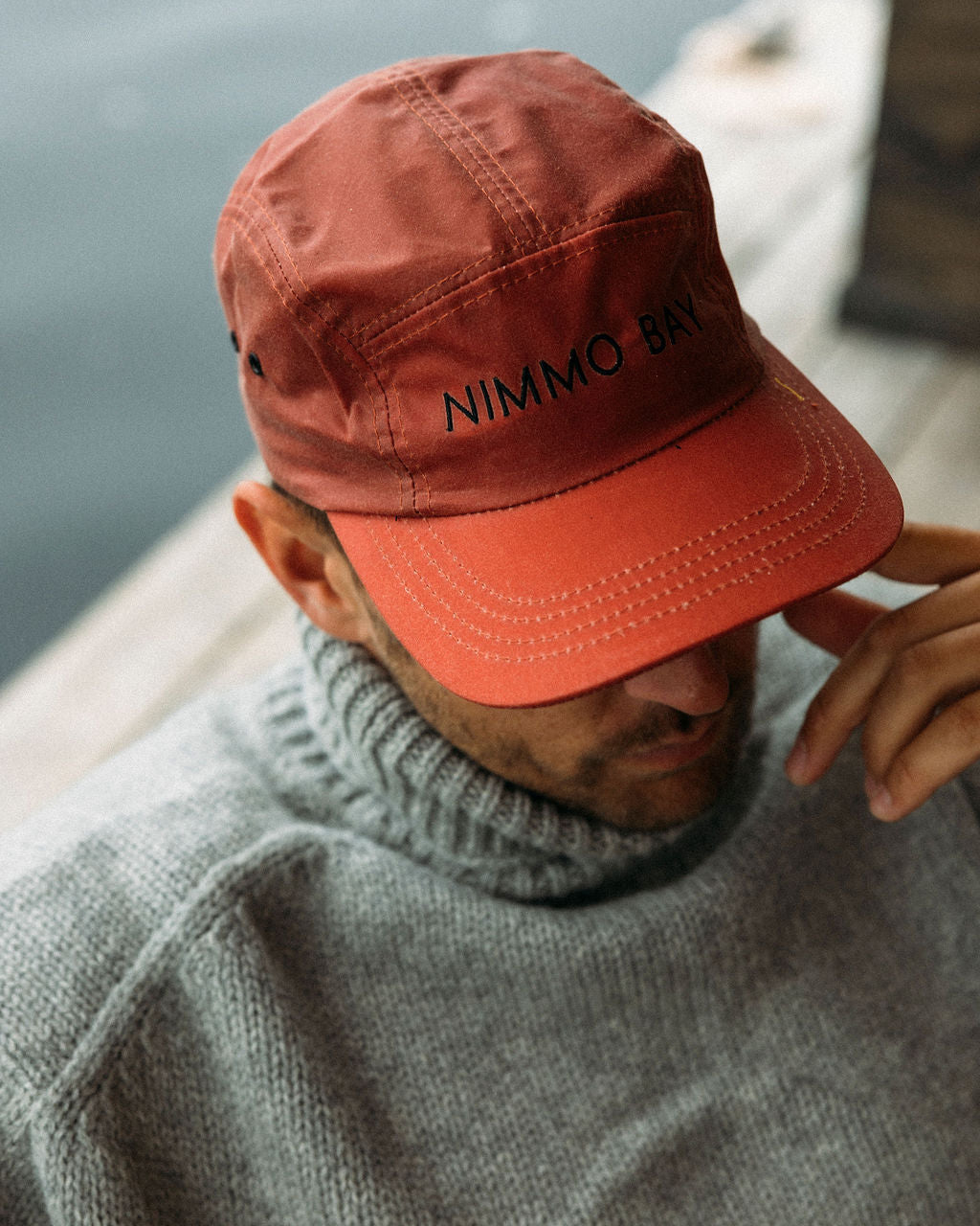 Person wearing a red anian tin cap with 'Nimmo Bay' branding, sitting by a body of water.