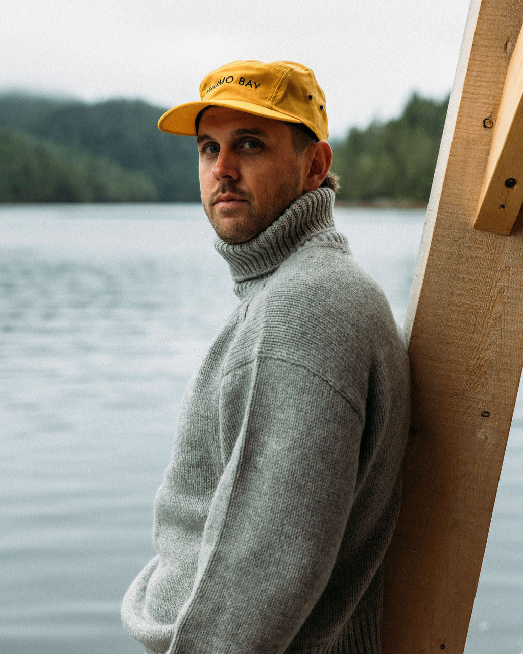 Man wearing a yellow anian tin cap and gray sweater by the ocean