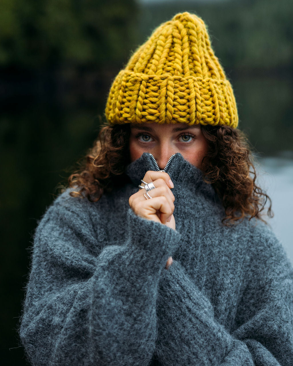 Person wearing a Ochre Diane Rudge knit toque and grey Bare Knitwear sweater standing against a forest background