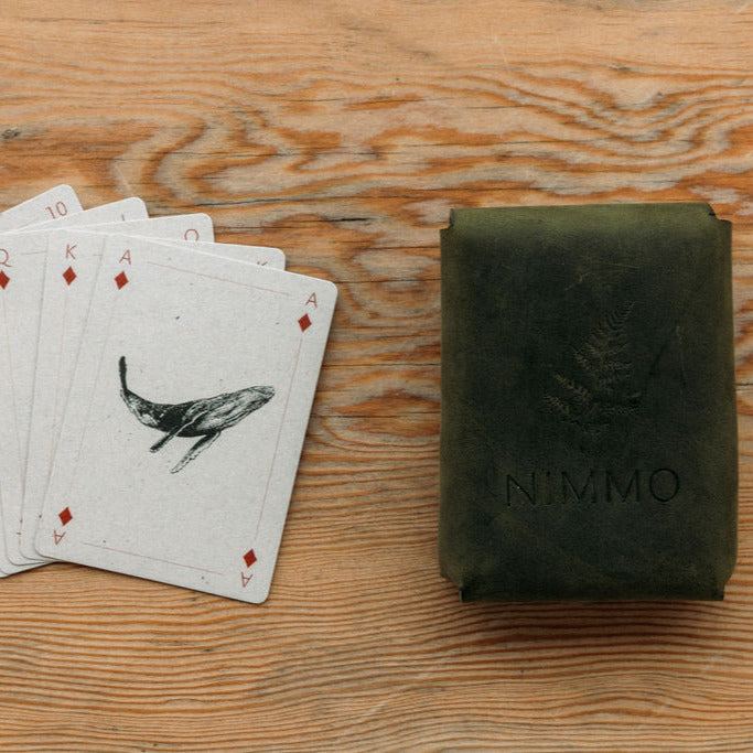 Playing cards with a whale illustration and a leather card case with 'Nimmo' branding on a wooden surface.