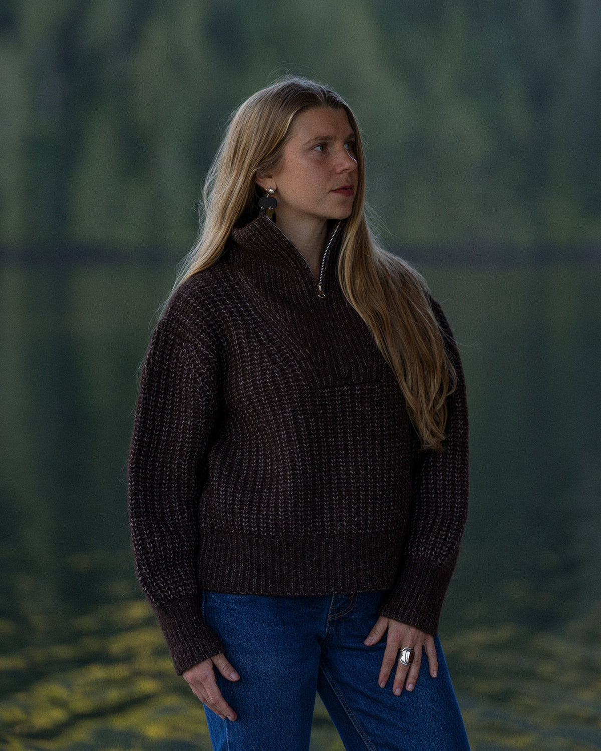 Woman wearing a Bare Knitwear carob brown quarter zip sweater with a forest background
