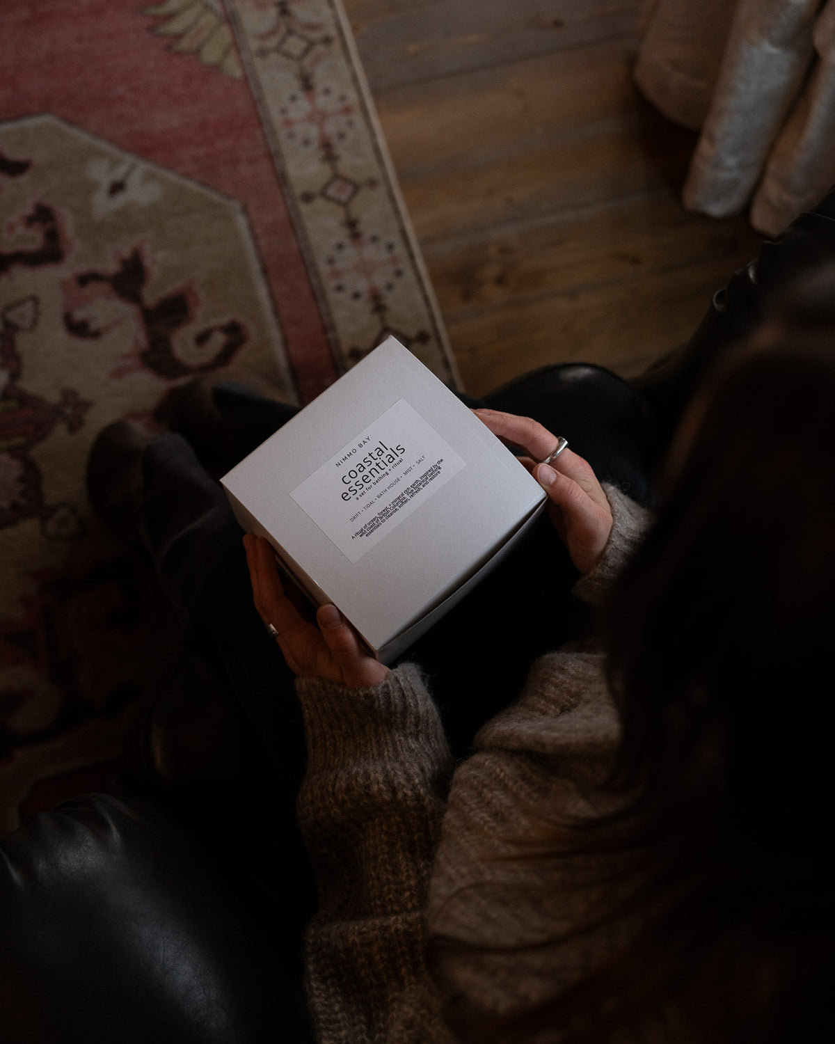 A person in a cozy indoor setting holds a white square box labeled 'coastal essentials".
