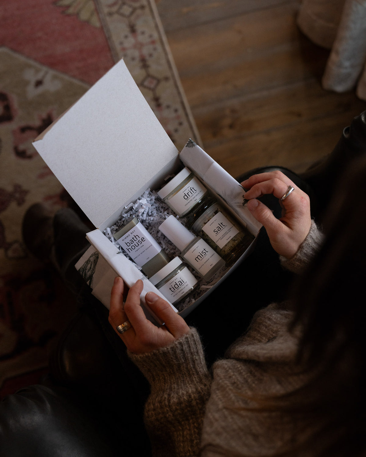 An open white gift box filled with skincare products, held by a person wearing a beige knit sweater. Visible products include a white tube labeled 'Mist' and several small jars with labels such as 'skin', 'face', and 'body'.
