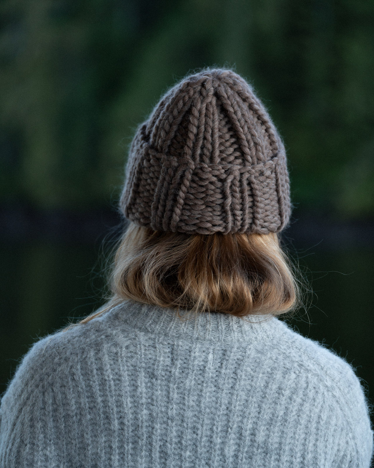 a woman from behind wearing a toque and knit sweater against a background of trees