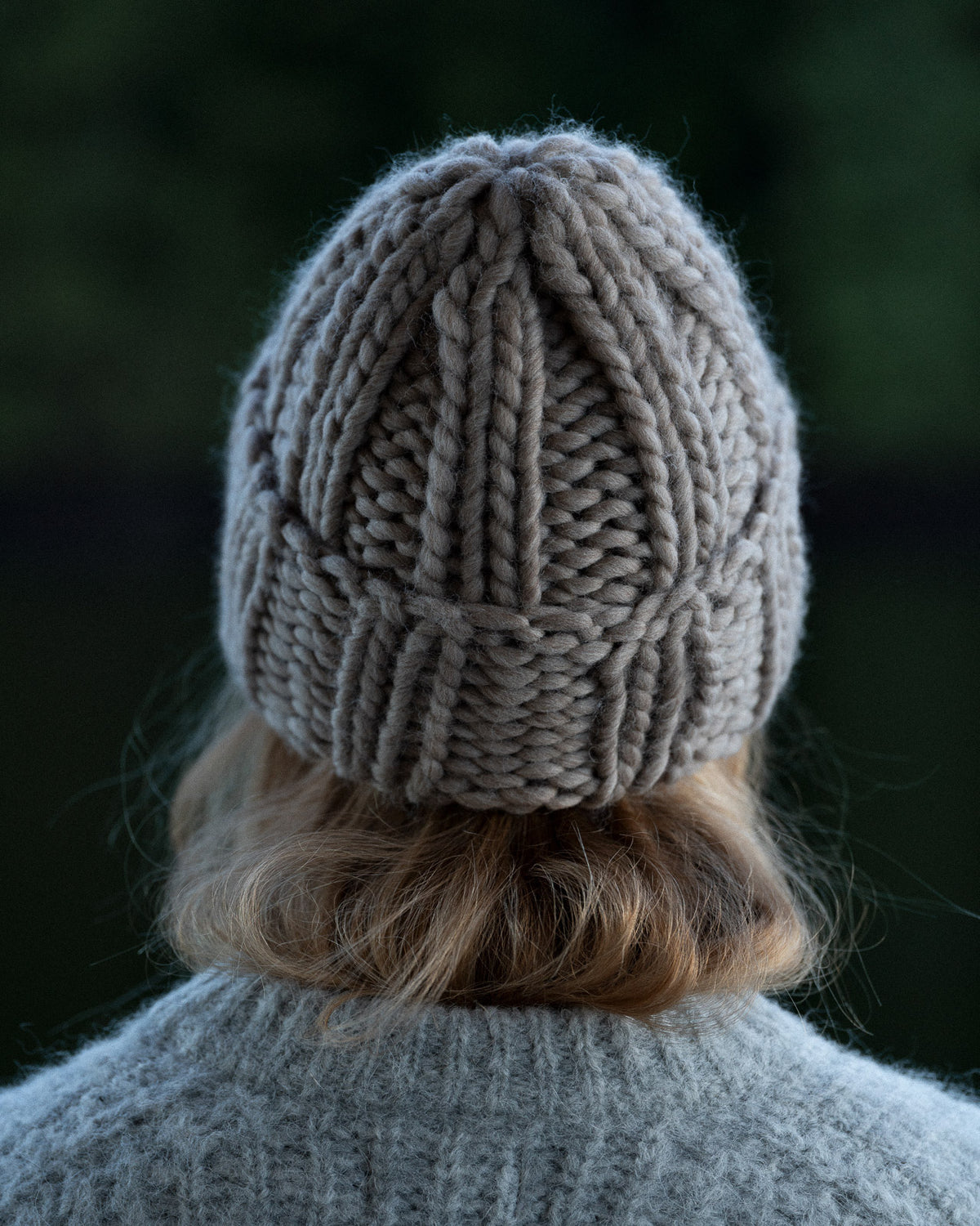 a woman from behind wearing a toque and knit sweater against a background of trees