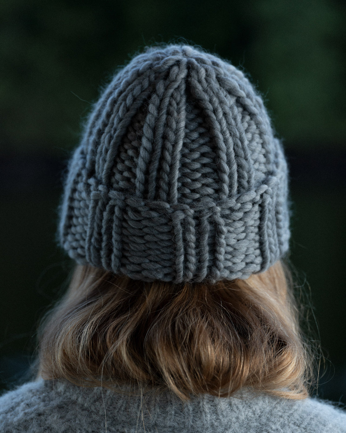 a woman from behind wearing a toque and knit sweater against a background of trees