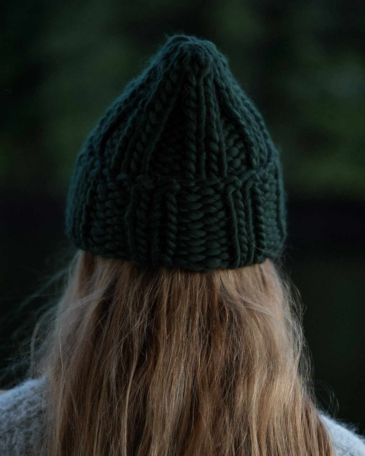 a woman from behind wearing a toque and knit sweater against a background of trees