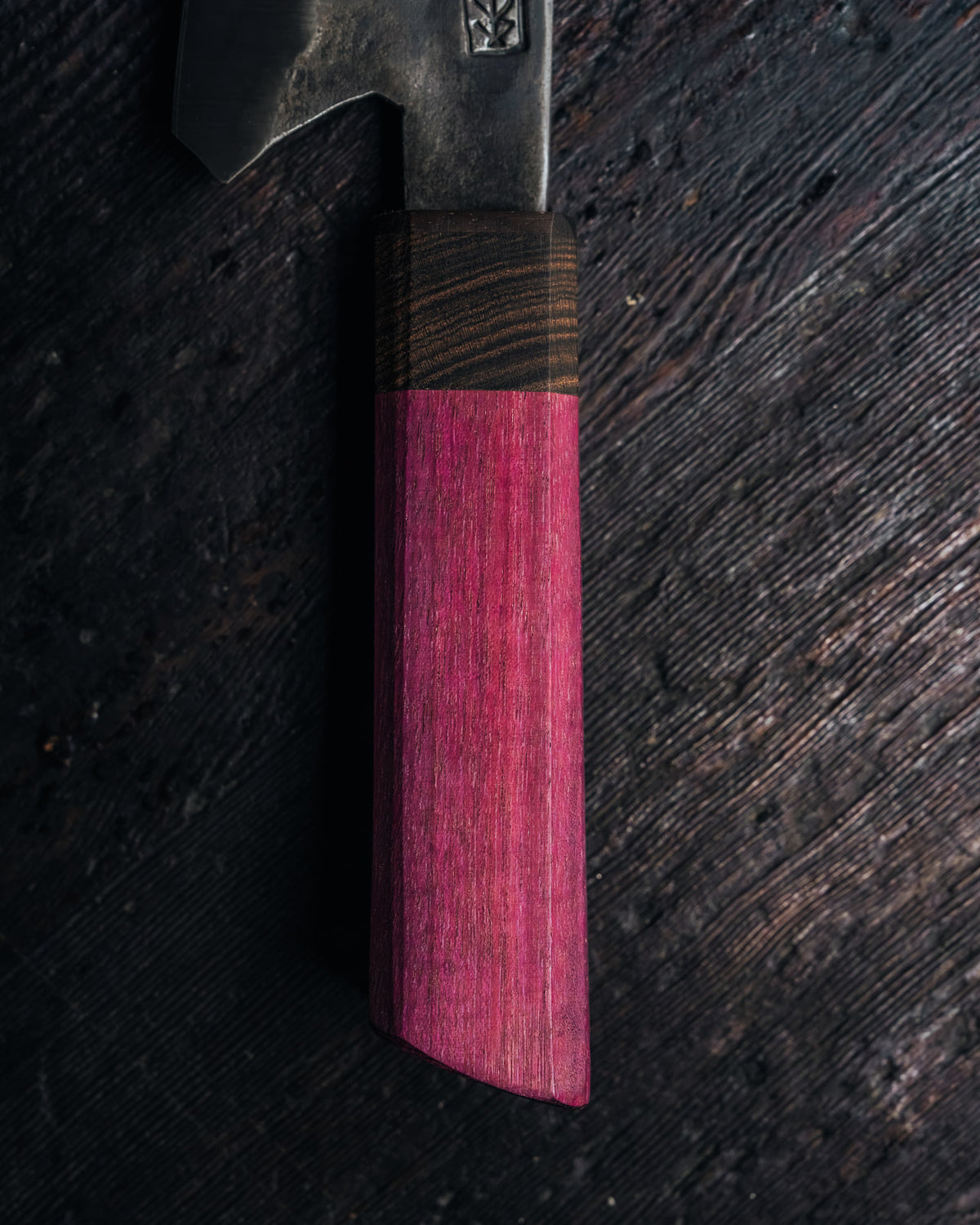 A close-up of a knife with a dark metal blade and a handle made of two distinct wood tones, resting on a dark, rustic wooden surface.