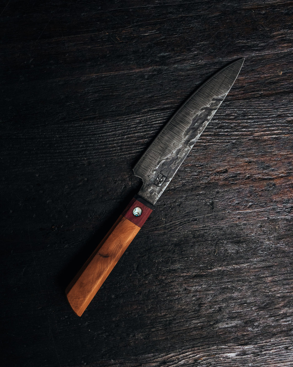 A Japanese-style kitchen knife with a dark patterned steel blade and a wooden handle, placed diagonally on a dark rustic wooden surface.