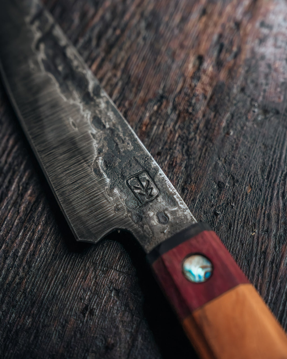 A close-up of a Japanese-style kitchen knife with a textured, hammered steel blade and a dual-tone wooden handle, resting on a dark rustic wooden surface.