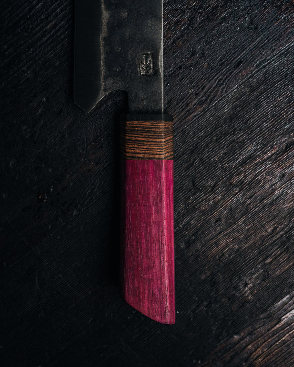 A close-up of a Japanese-style kitchen knife with a dark, textured steel blade and a vibrant magenta-pink wooden handle with a striped wood collar, set against a dark, rustic wooden background.