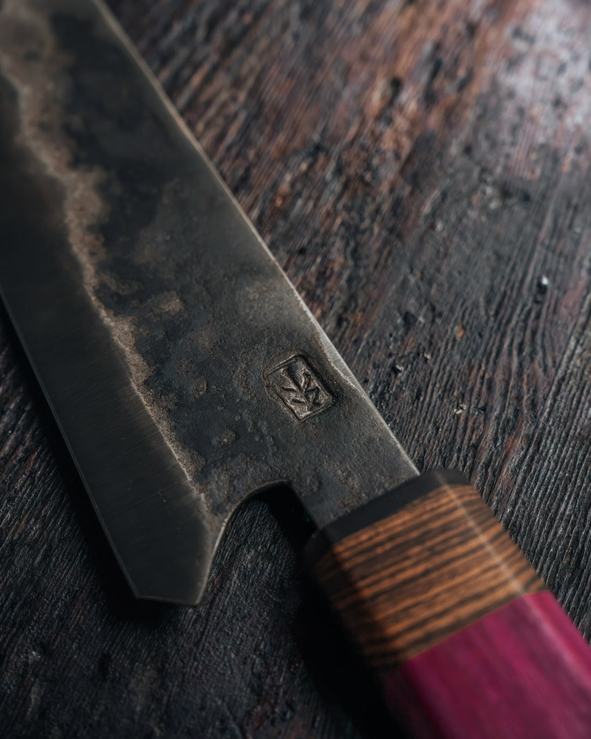 A close-up of a Japanese-style kitchen knife with a dark, textured forged steel blade and a handle featuring a brown wood grain section and a magenta-pink end, resting on a dark rustic wooden surface.