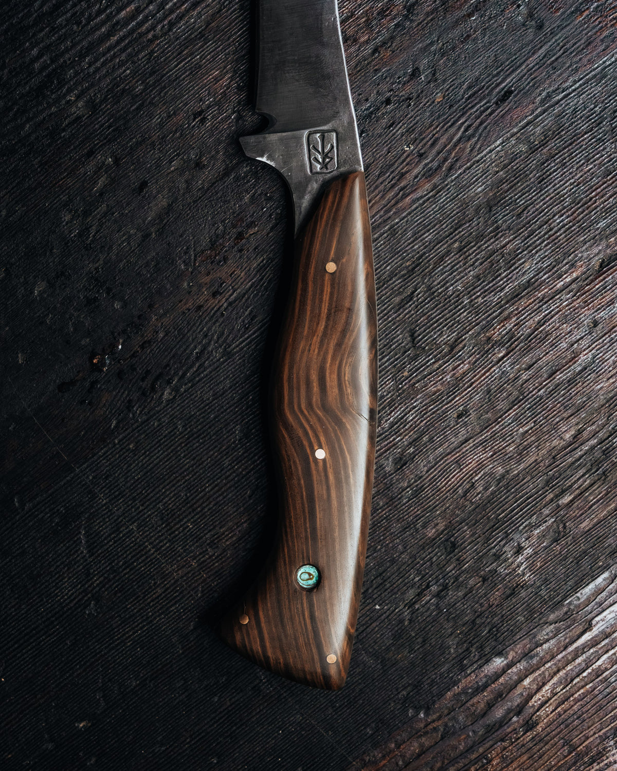 A fixed-blade knife with a dark wood handle and a dark metal blade, laid diagonally on a dark textured wooden surface.