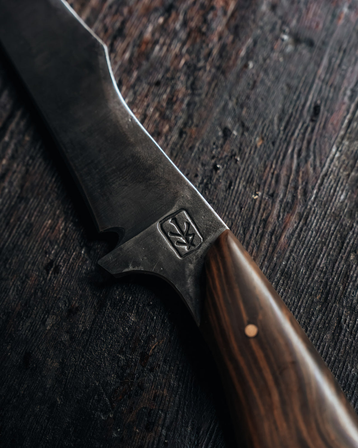 A close-up of a fixed-blade knife with a dark, textured steel blade and a polished brown wood handle, resting on a dark rustic wooden surface. The blade features an embossed logo.