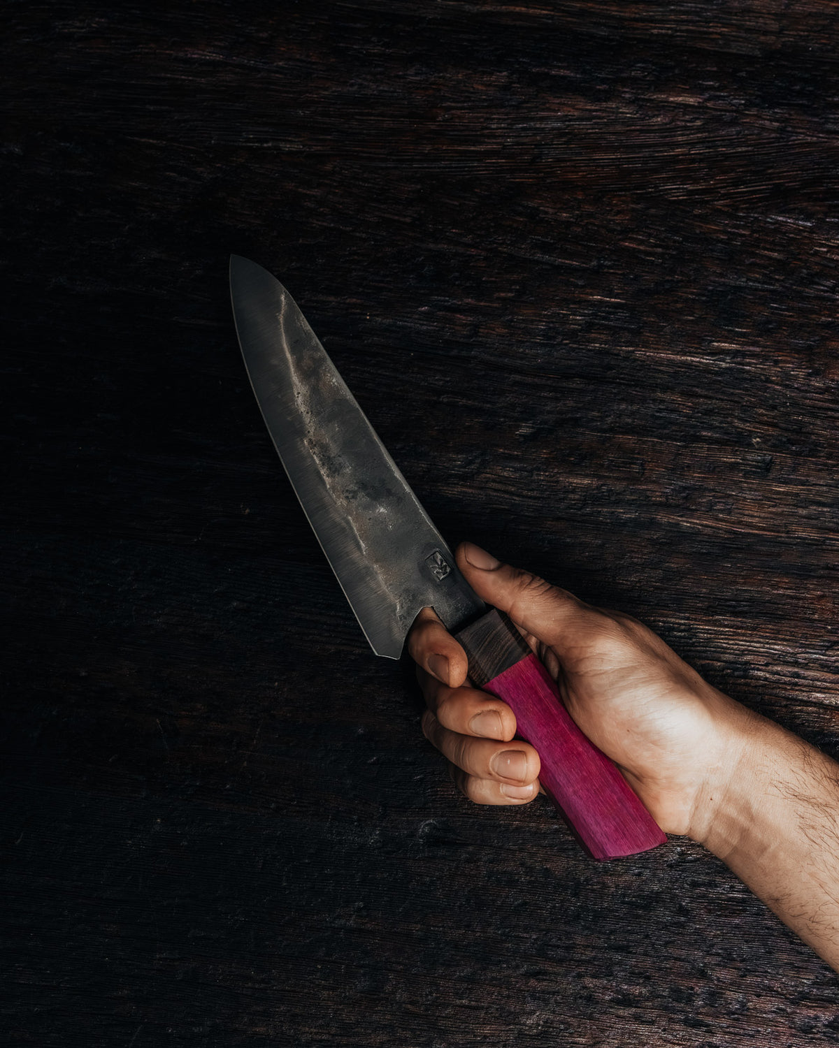 A hand holding a large fixed-blade knife with a dark steel blade and a magenta-pink handle, set against a dark, rustic wooden background.