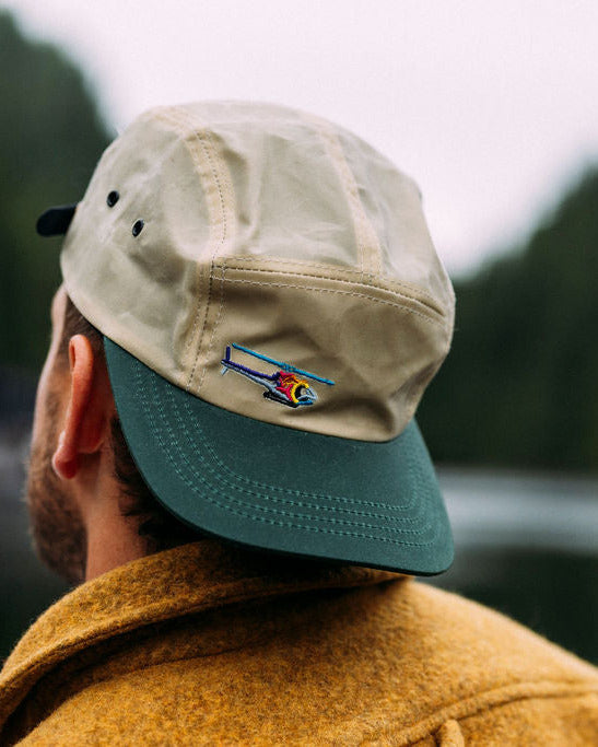 a man wearing a legacy anian tin cap with a helifish embroidery in front of a wooded background