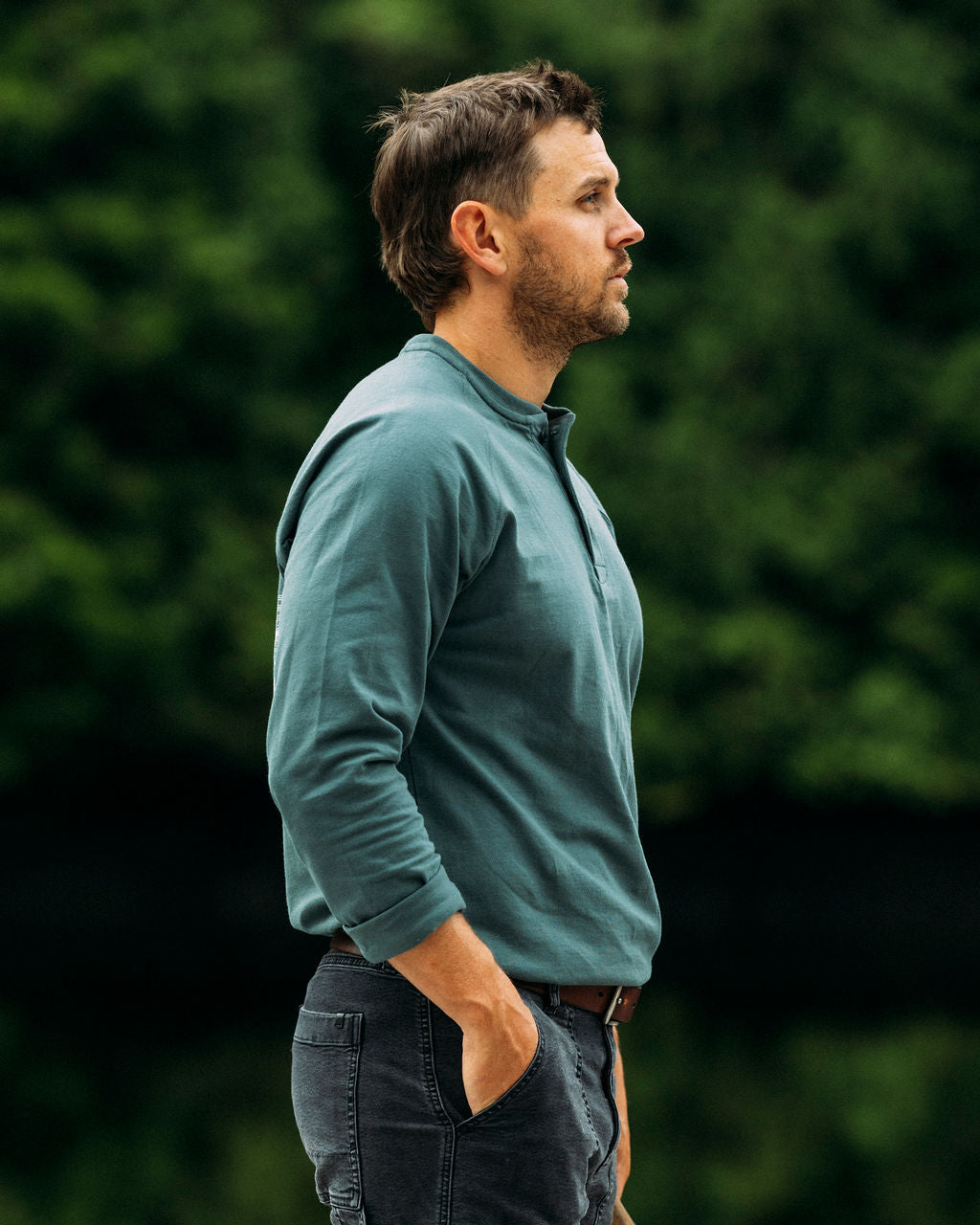 Man wearing a green anian henley standing in a forest.