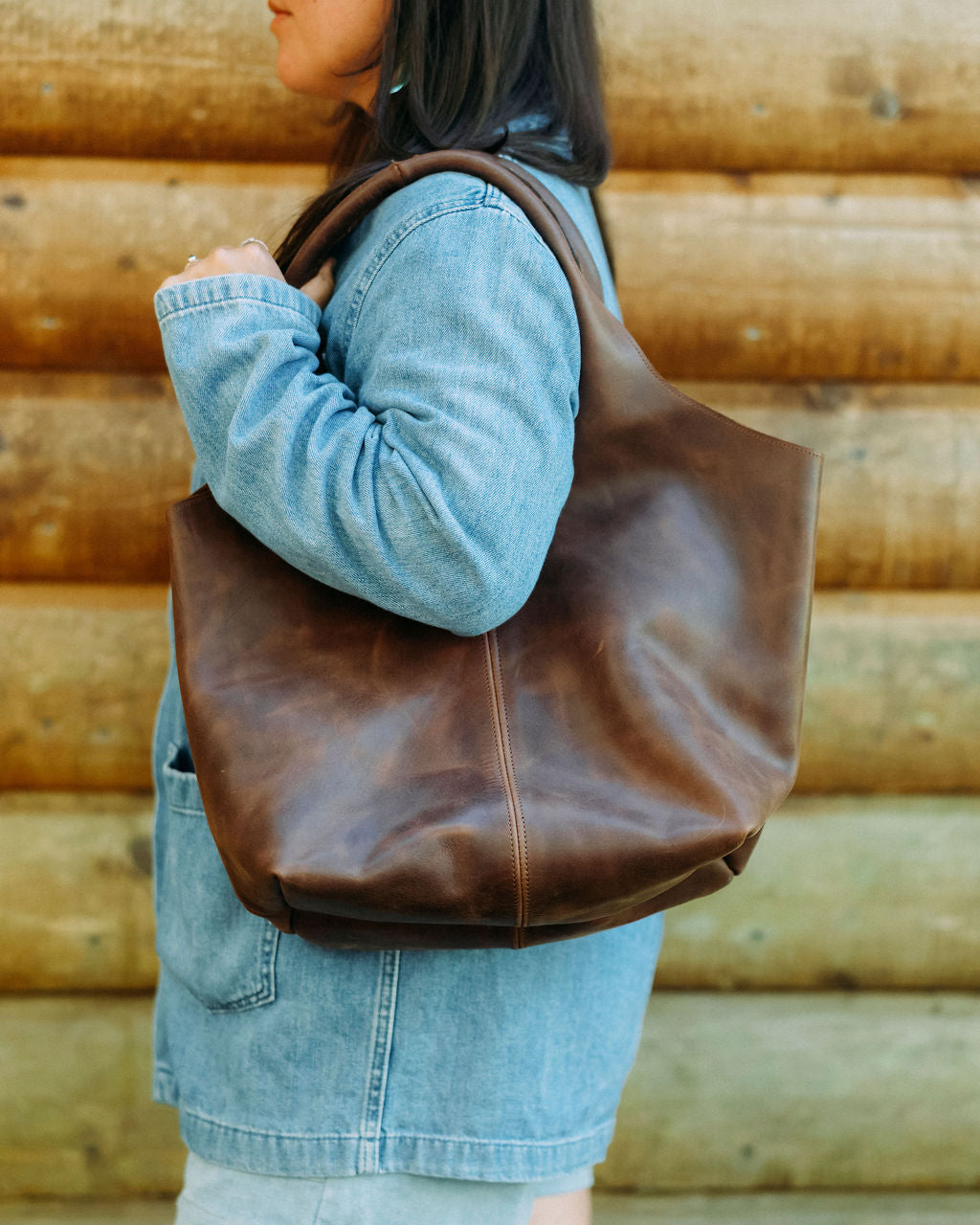 Person wearing a brown leather tote bag against a wooden wall.