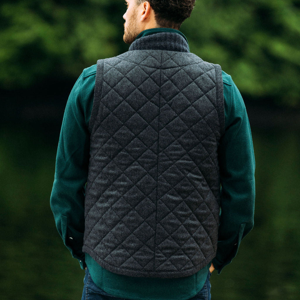 Man wearing an Anian quilted wool vest over a green modern melton shirt with a blurred forest background