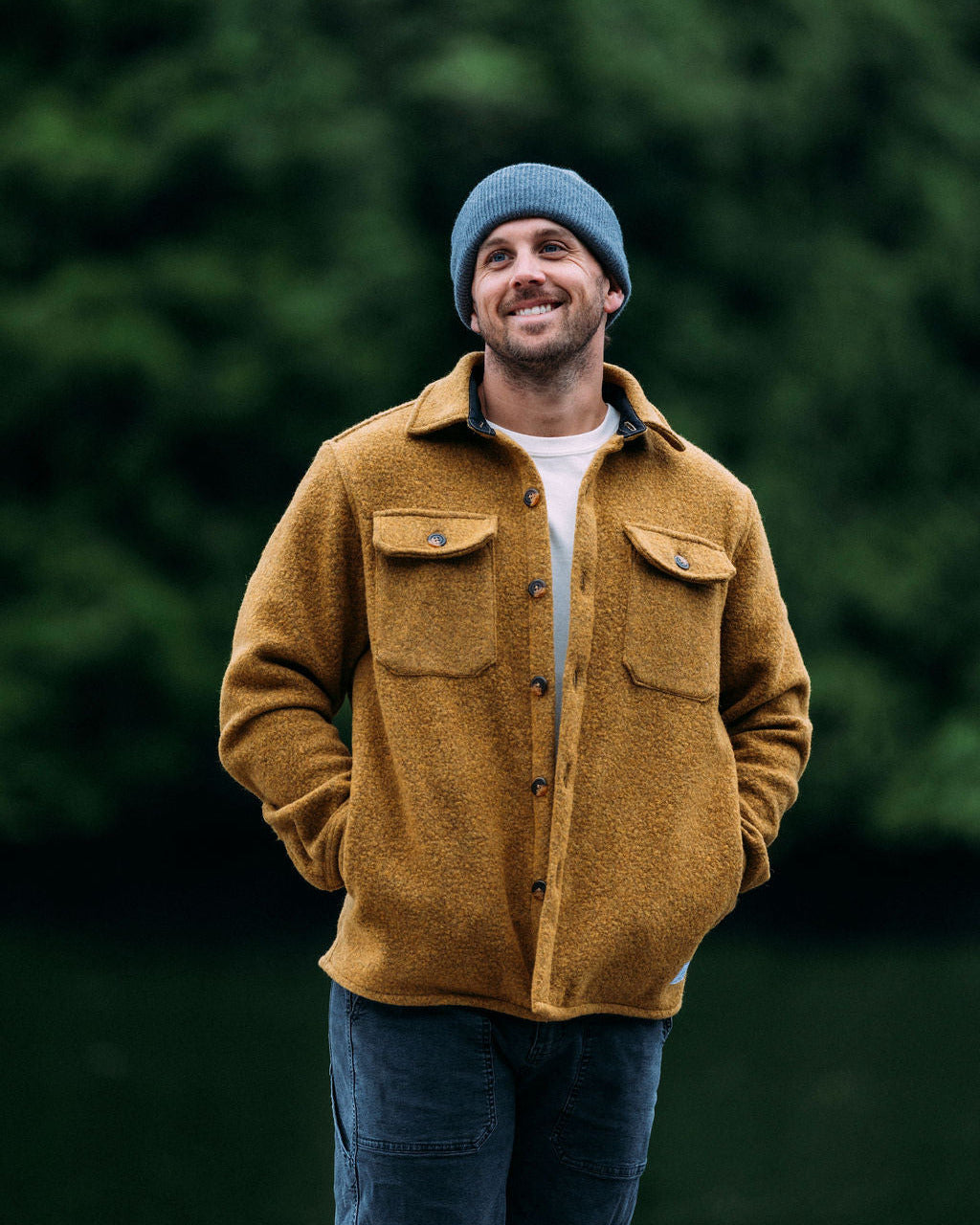 Man wearing a mustard yellow jacket and blue beanie standing in front of green trees