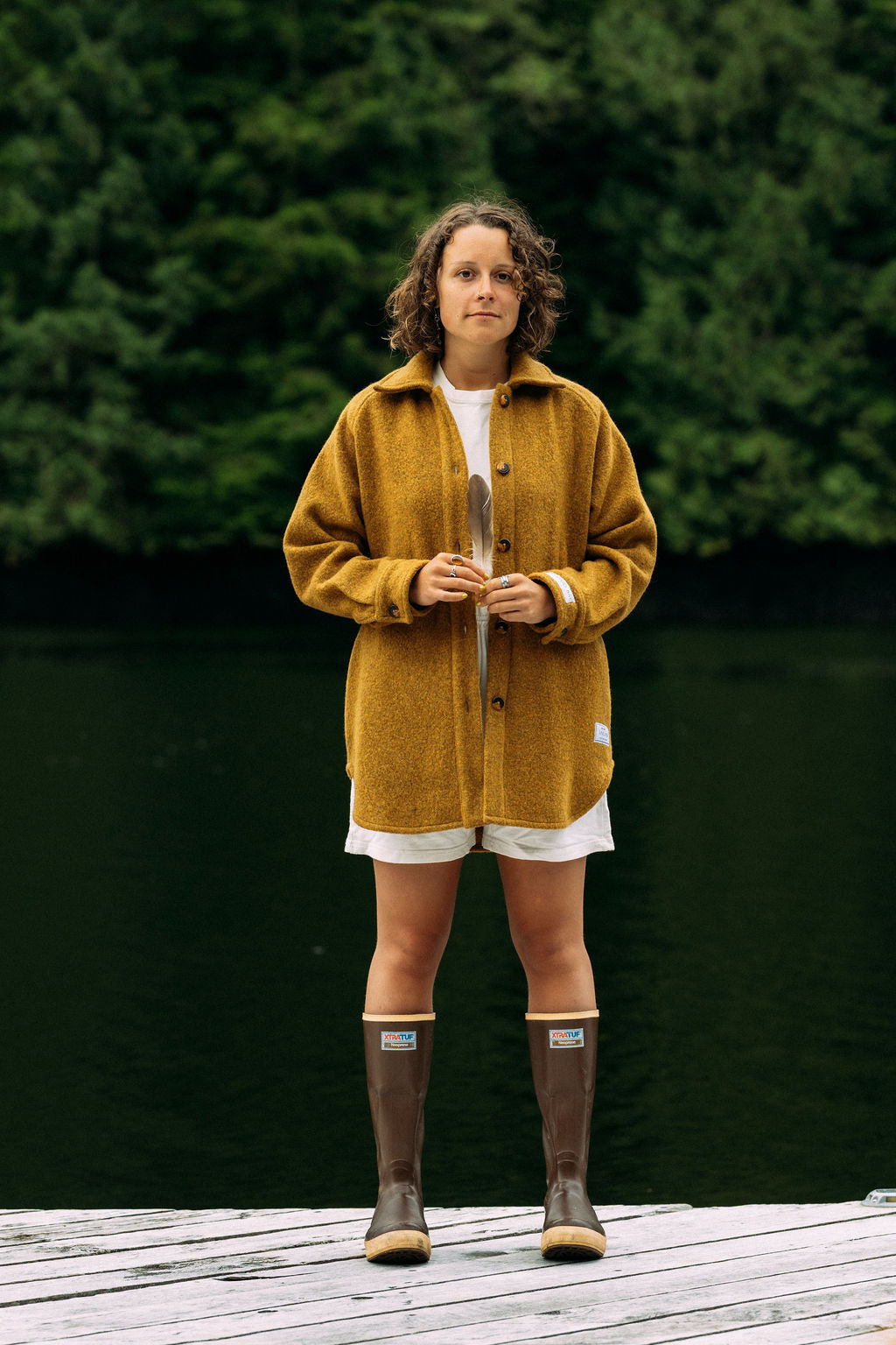 Person wearing a mustard yellow jacket and brown boots standing on a wooden deck with greenery in the background