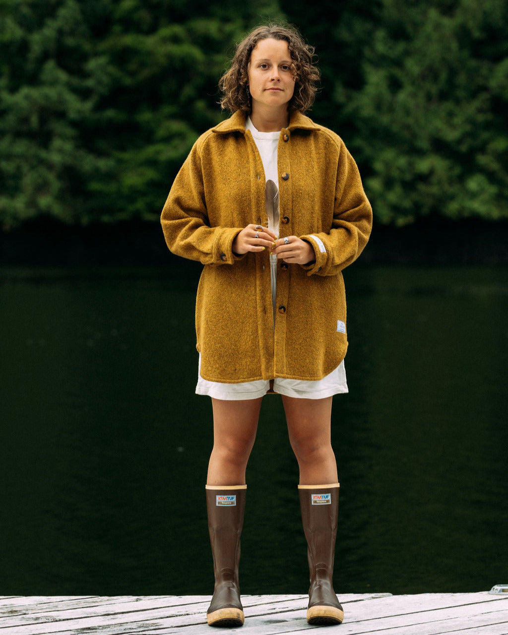 Person wearing a gold anian loft coat and brown boots standing on a wooden deck with greenery in the background