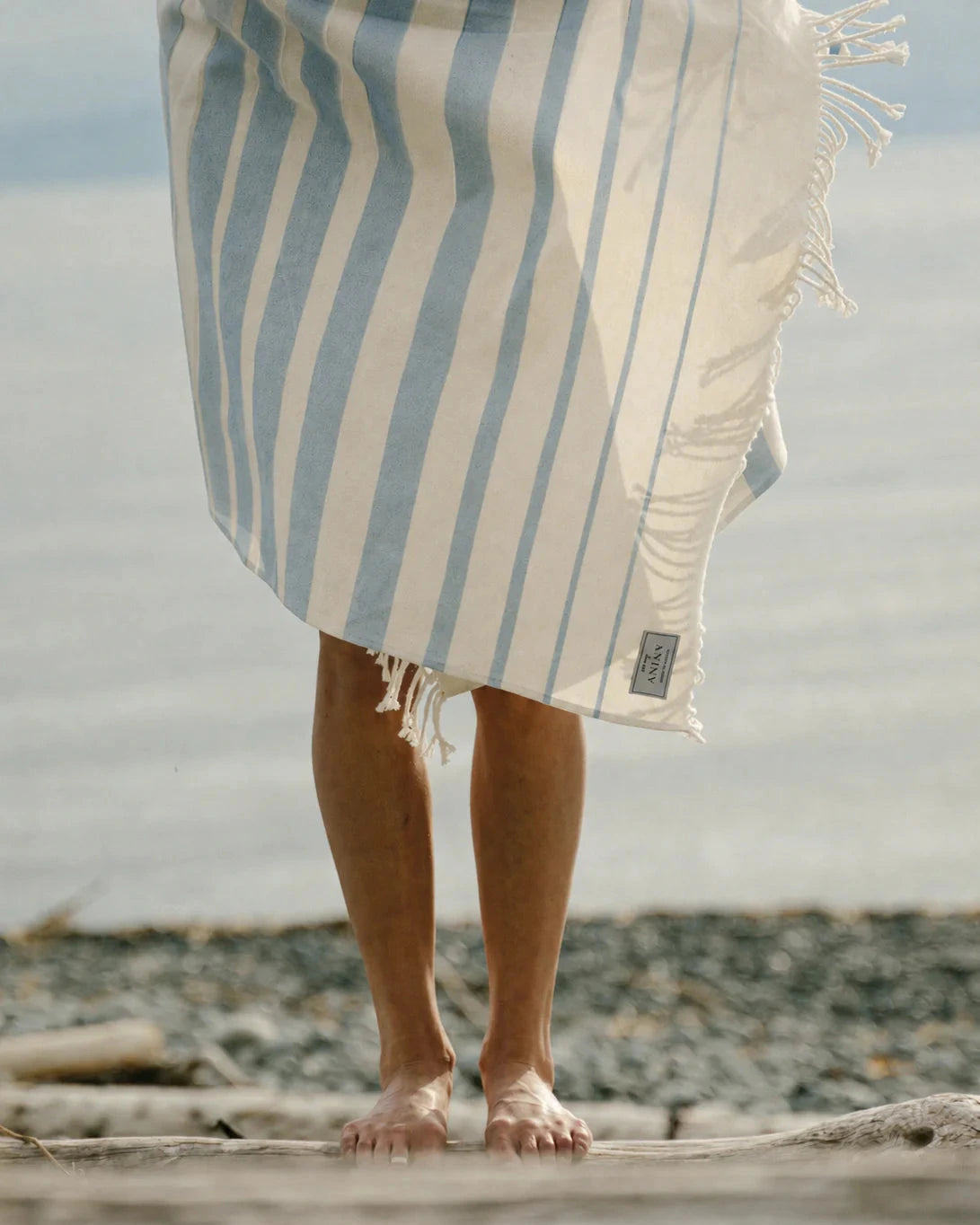 Person standing on a log by the water with a striped towel draped over their shoulders.