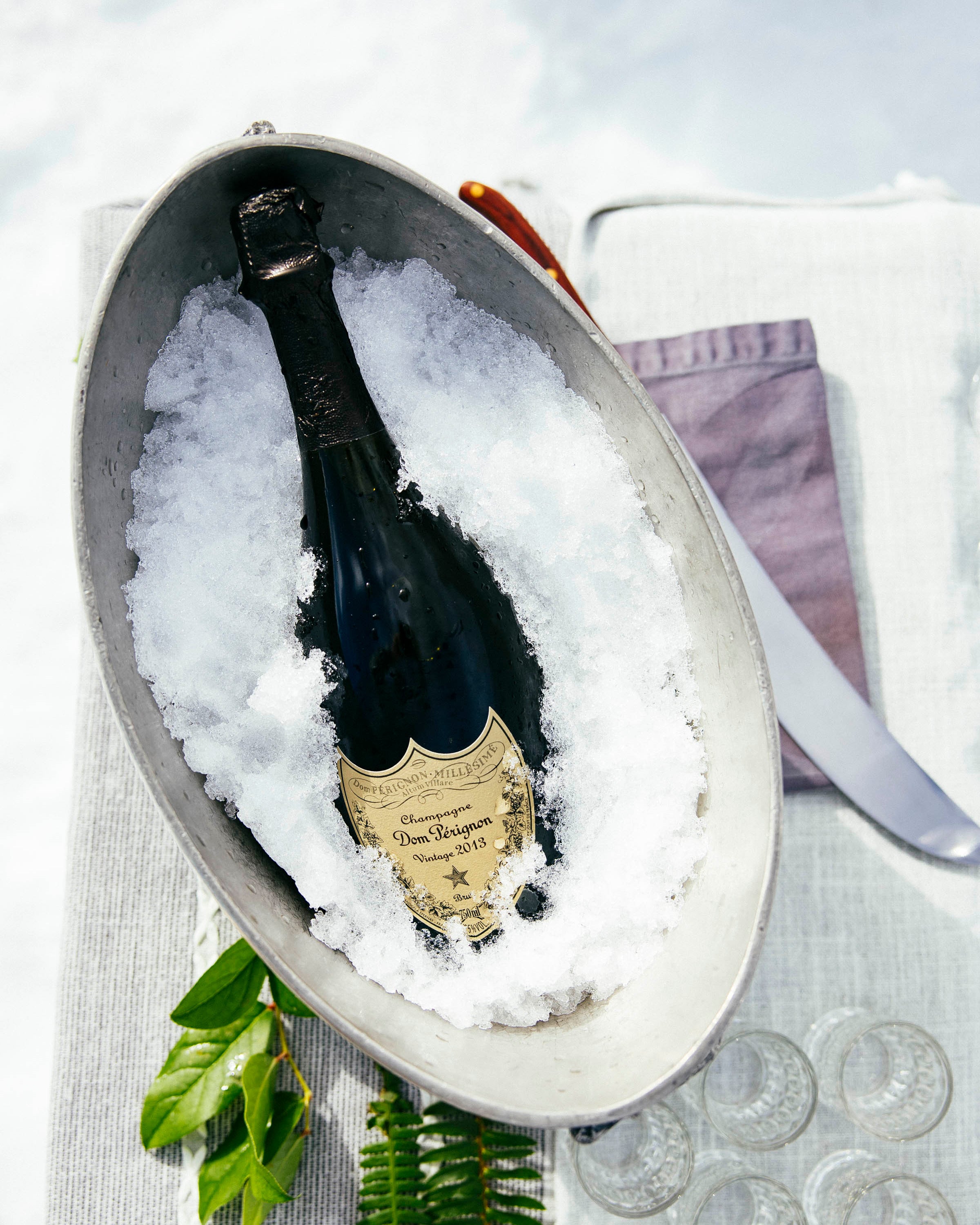 Bottle of champagne in a metal ice bucket with ice, surrounded by glasses on a white tablecloth.