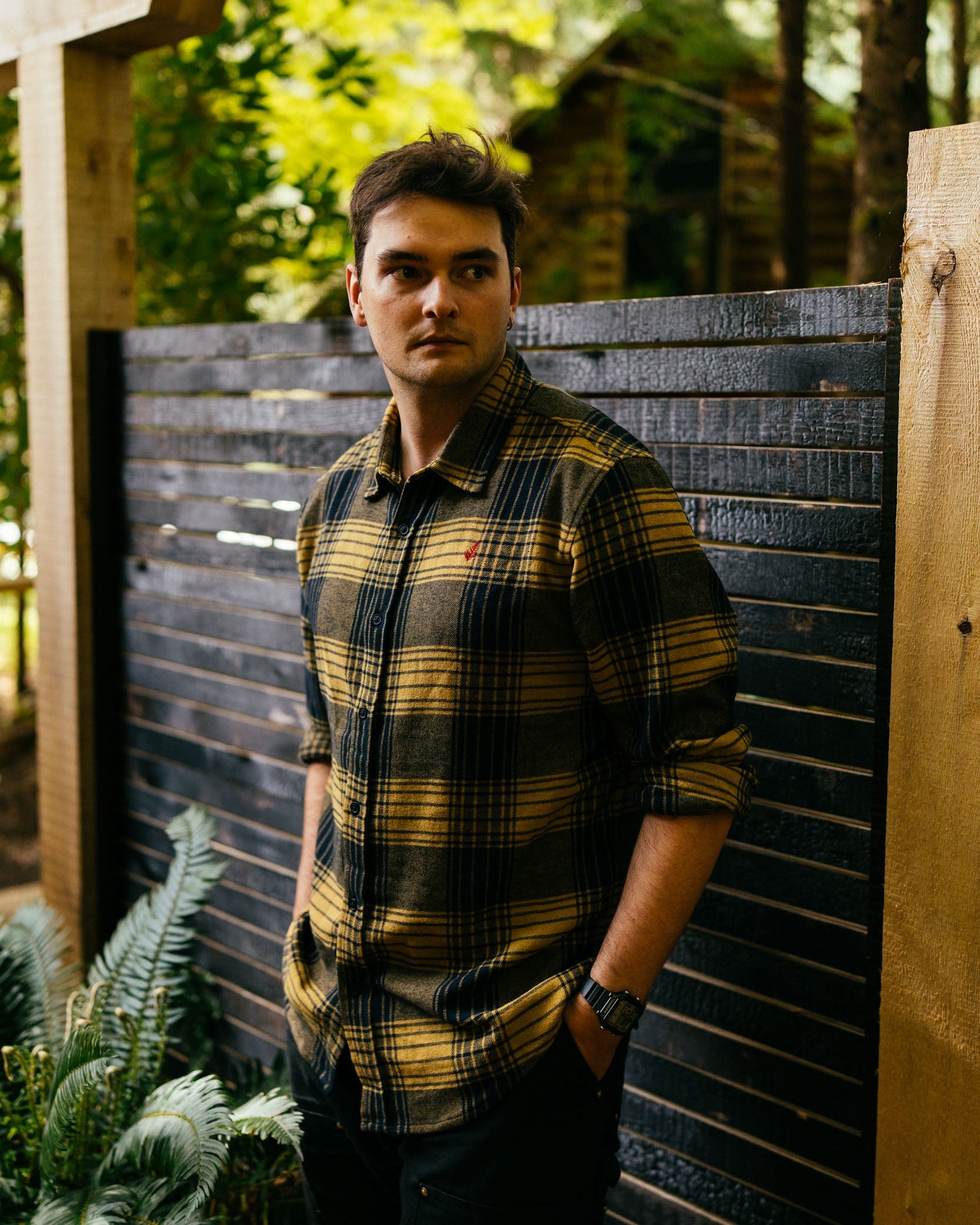 Man wearing a plaid shirt standing in front of a wooden fence with greenery in the background