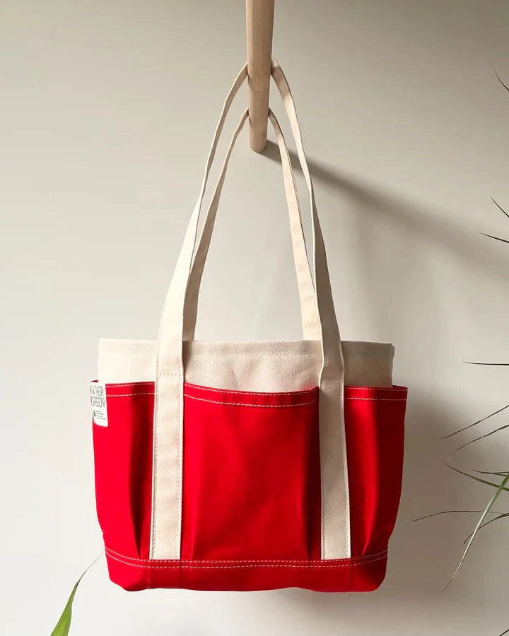 Red Rather green amiga petite tote bag hanging on a hook against a neutral wall with a plant in the corner.