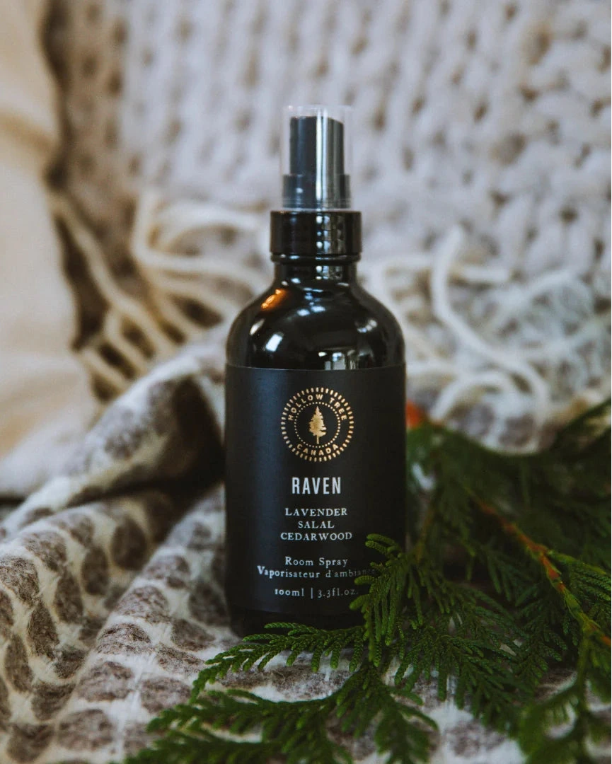 A black glass room spray bottle labeled 'RAVEN Lavender, Salal, Cedarwood' sits on a cozy beige knit blanket with green pine branches around it.