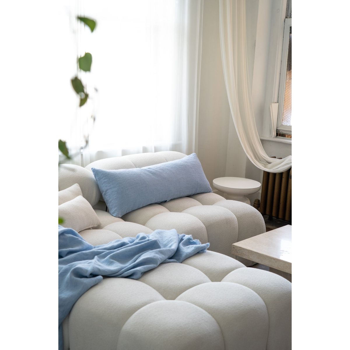 Beige sofa with blue cushions and a blanket in a room with white curtains and a plant.