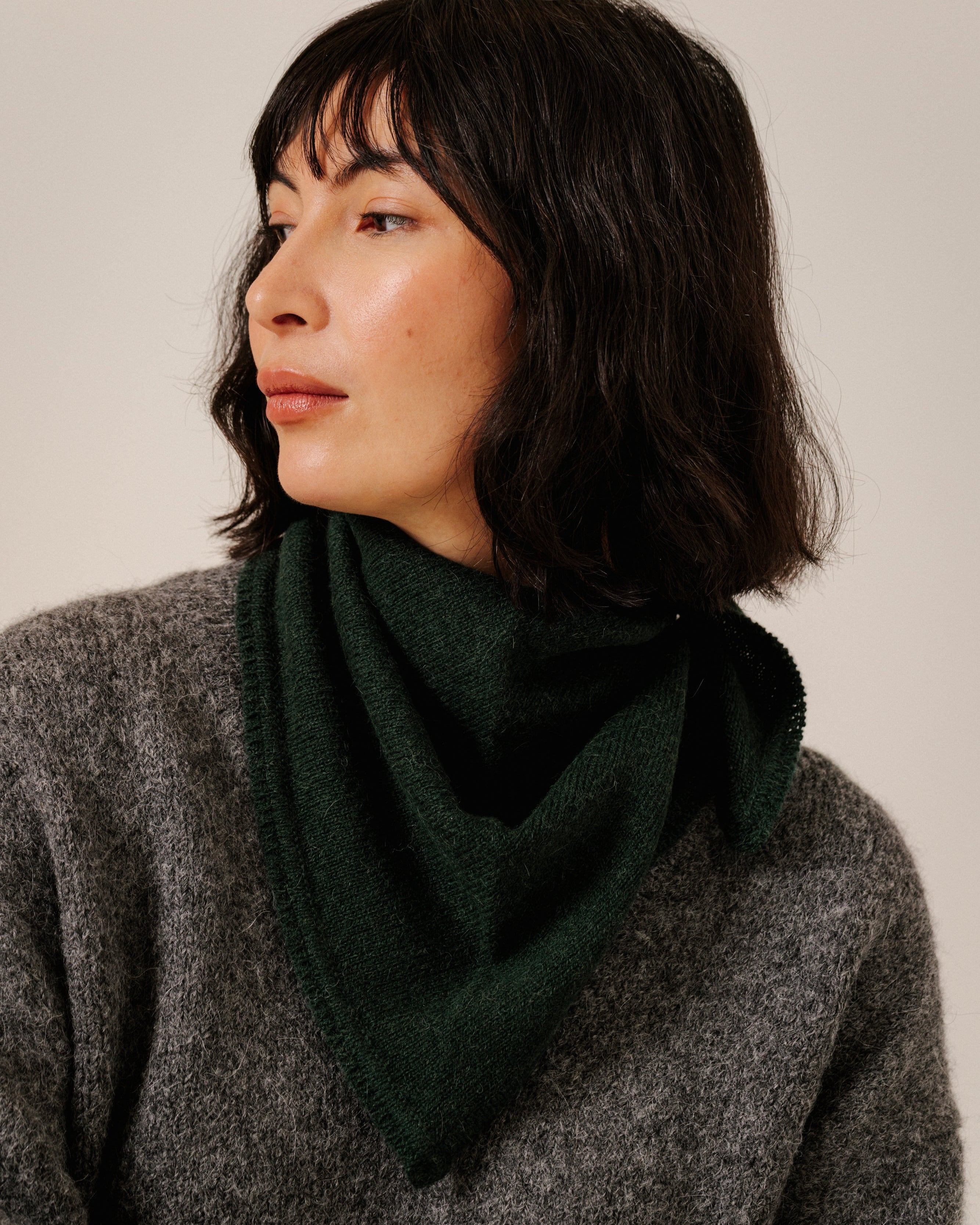 A woman with short dark hair wearing a dark forest green cowl scarf and a textured gray knit sweater, photographed against a plain light beige background.