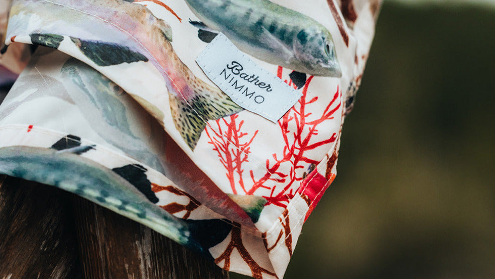 A close-up of a lightweight fabric accessory draped over a rustic wooden post, featuring a detailed print of fish and coral on a white background. A small white label with the brand name 'Breezin' NINJA' is visible.
