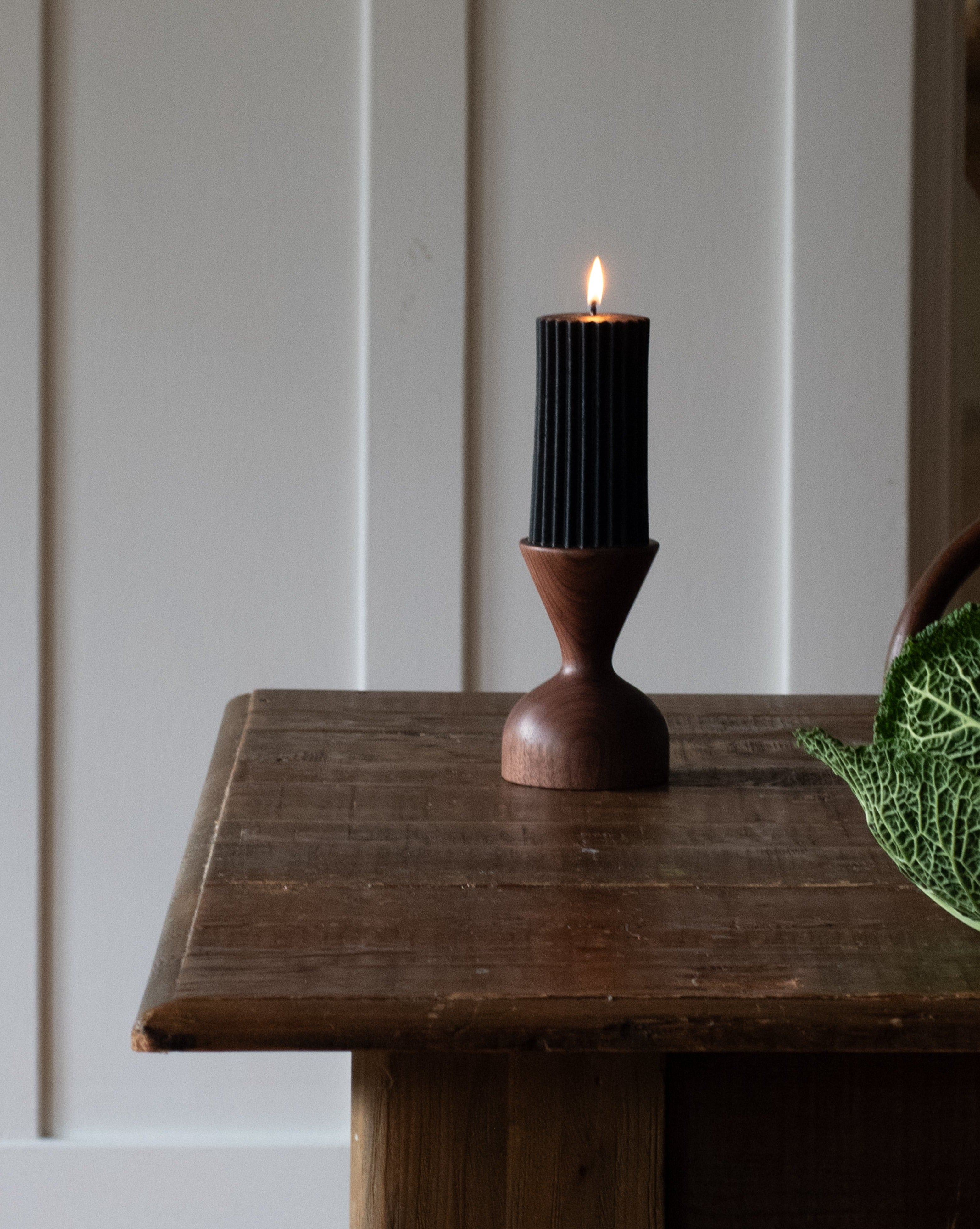 A brown wooden pillar candle holder by Miller + Co. is displayed on a wooden table with a lit black pillar candle in it, against a white panelled wall.