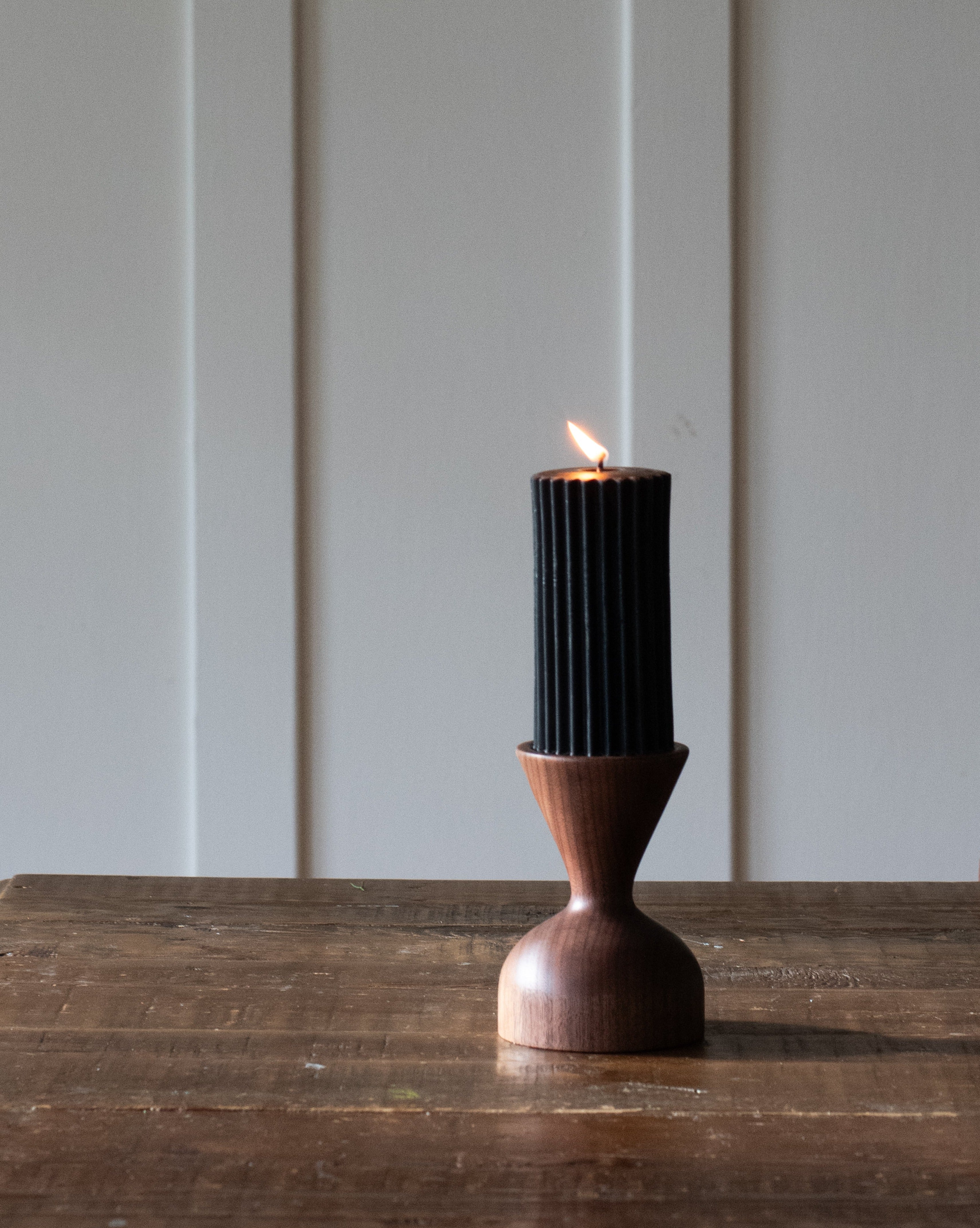A brown wooden pillar candle holder by Miller + Co. is displayed on a wooden table with a lit black pillar candle in it, against a white panelled wall.