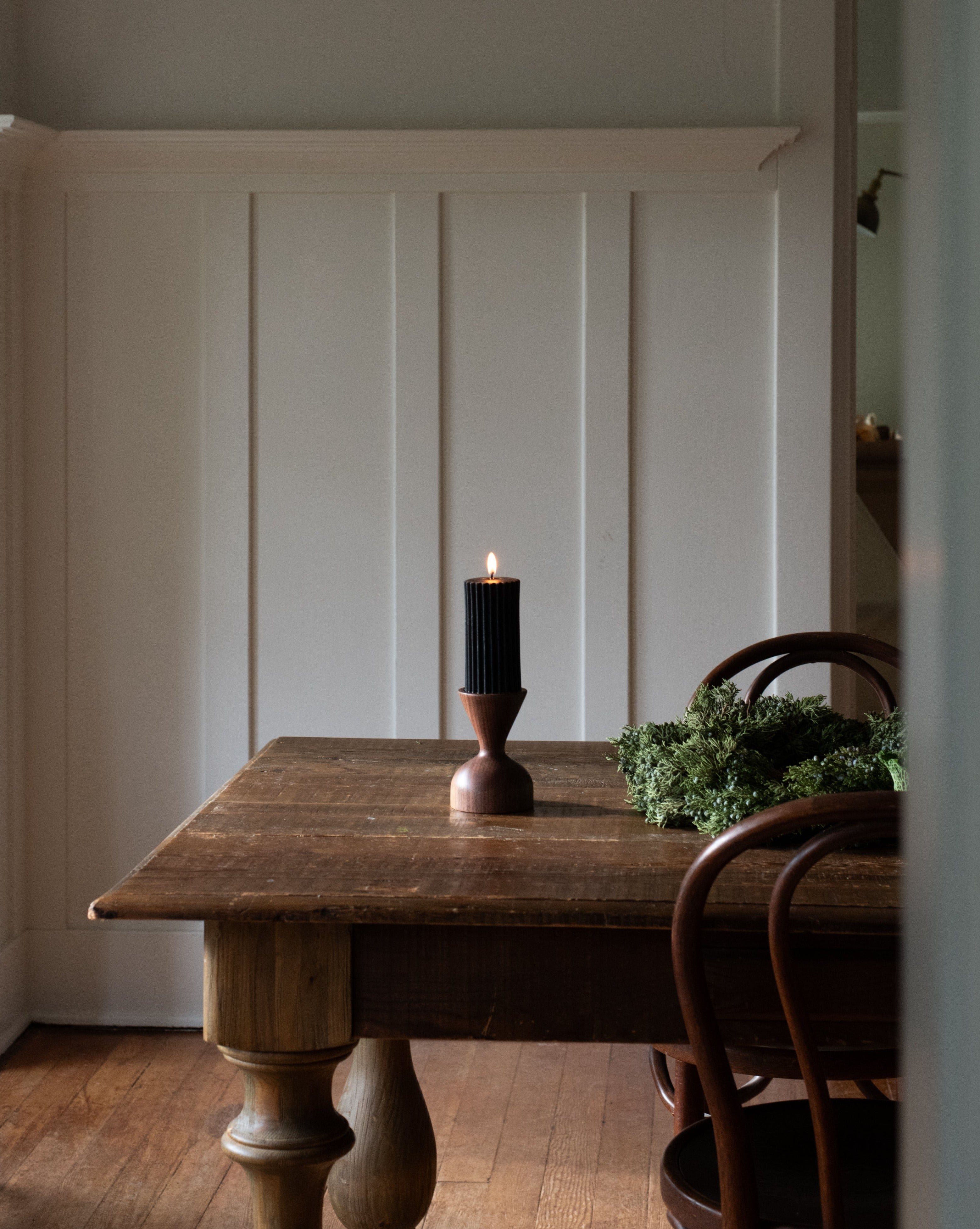 A brown wooden pillar candle holder by Miller + Co. is displayed on a wooden table with a lit black pillar candle in it, against a white panelled wall.