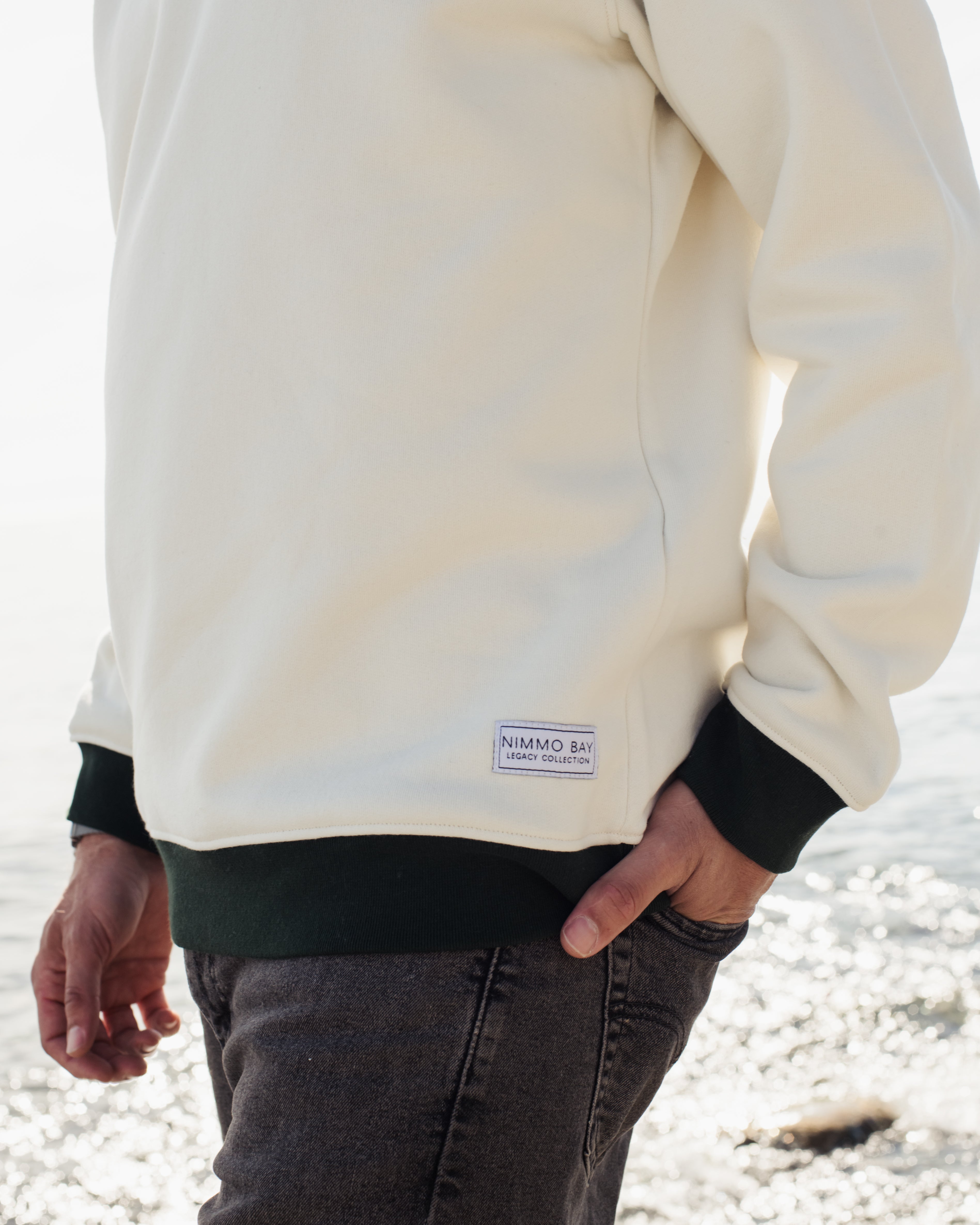 a man wearing the legacy crewneck sweater from anian in front of the ocean. the nimmo bay tag detail is shown
