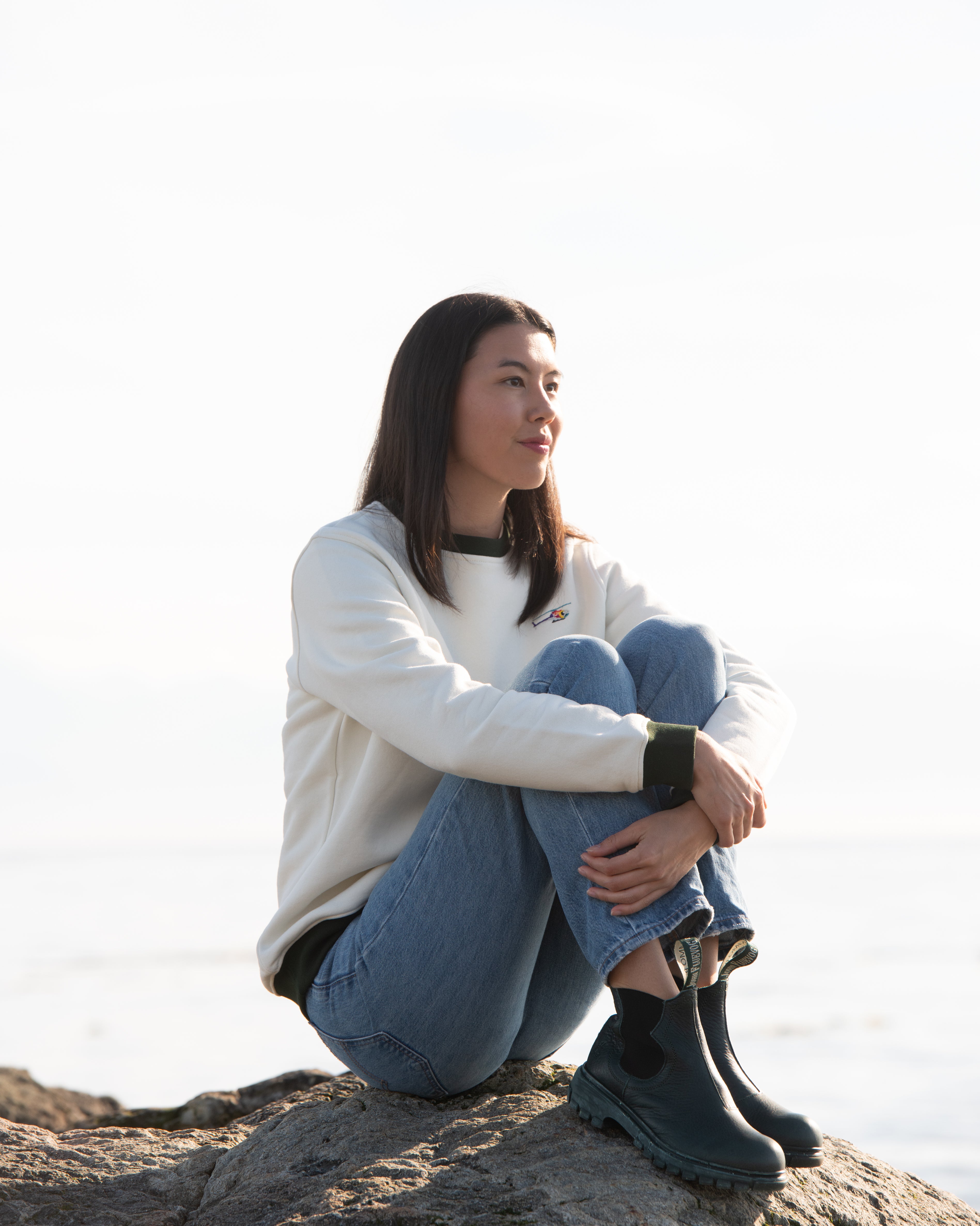 A person sitting on a rock wearing a cream-colored crewneck sweatshirt with green cuffs and a logo on the chest, paired with blue jeans and black shoes.