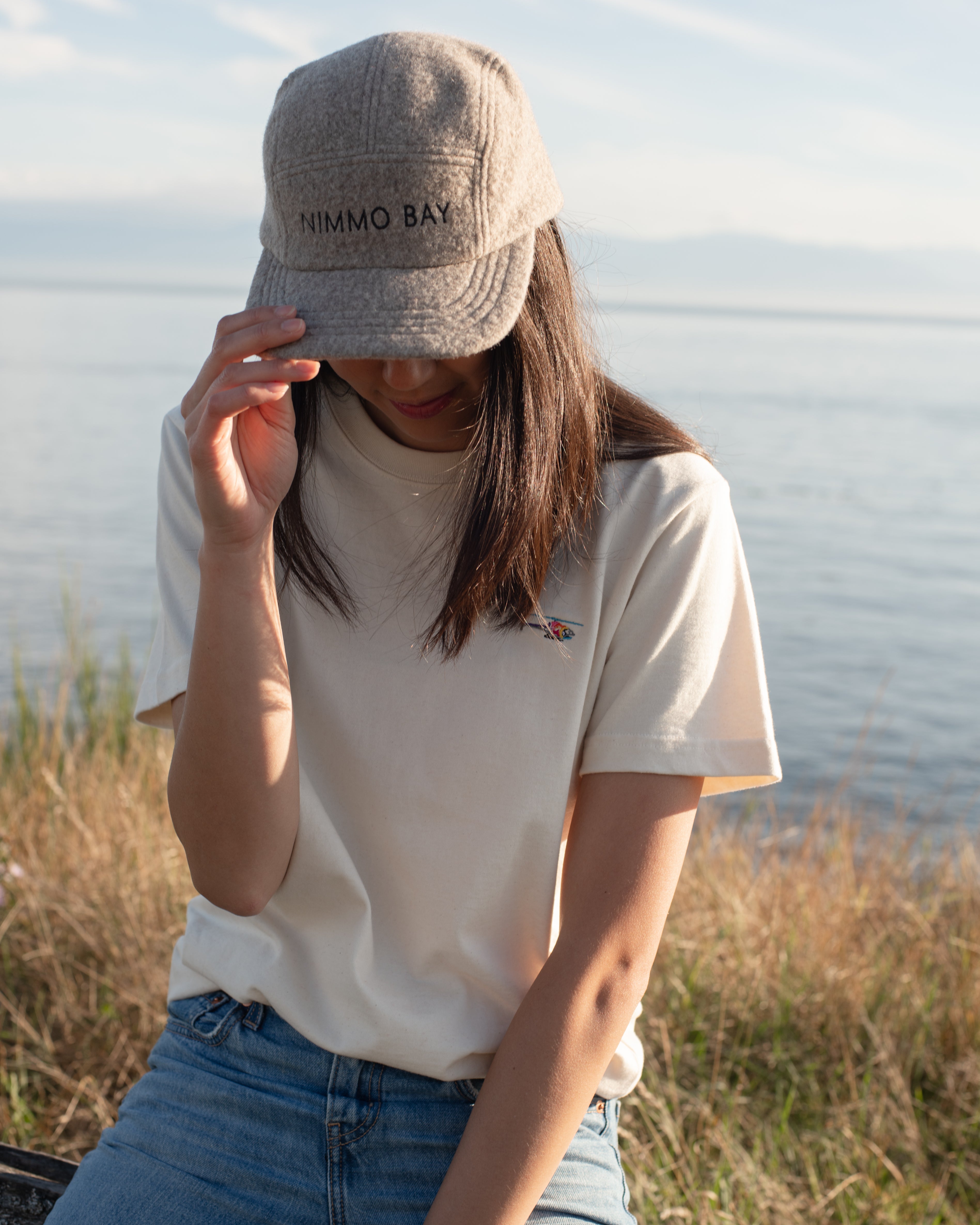 A person wearing a beige anian fuzzy cap with the Nimmo Bay logo, an antique leather buckle, and an adjustable leather strap.