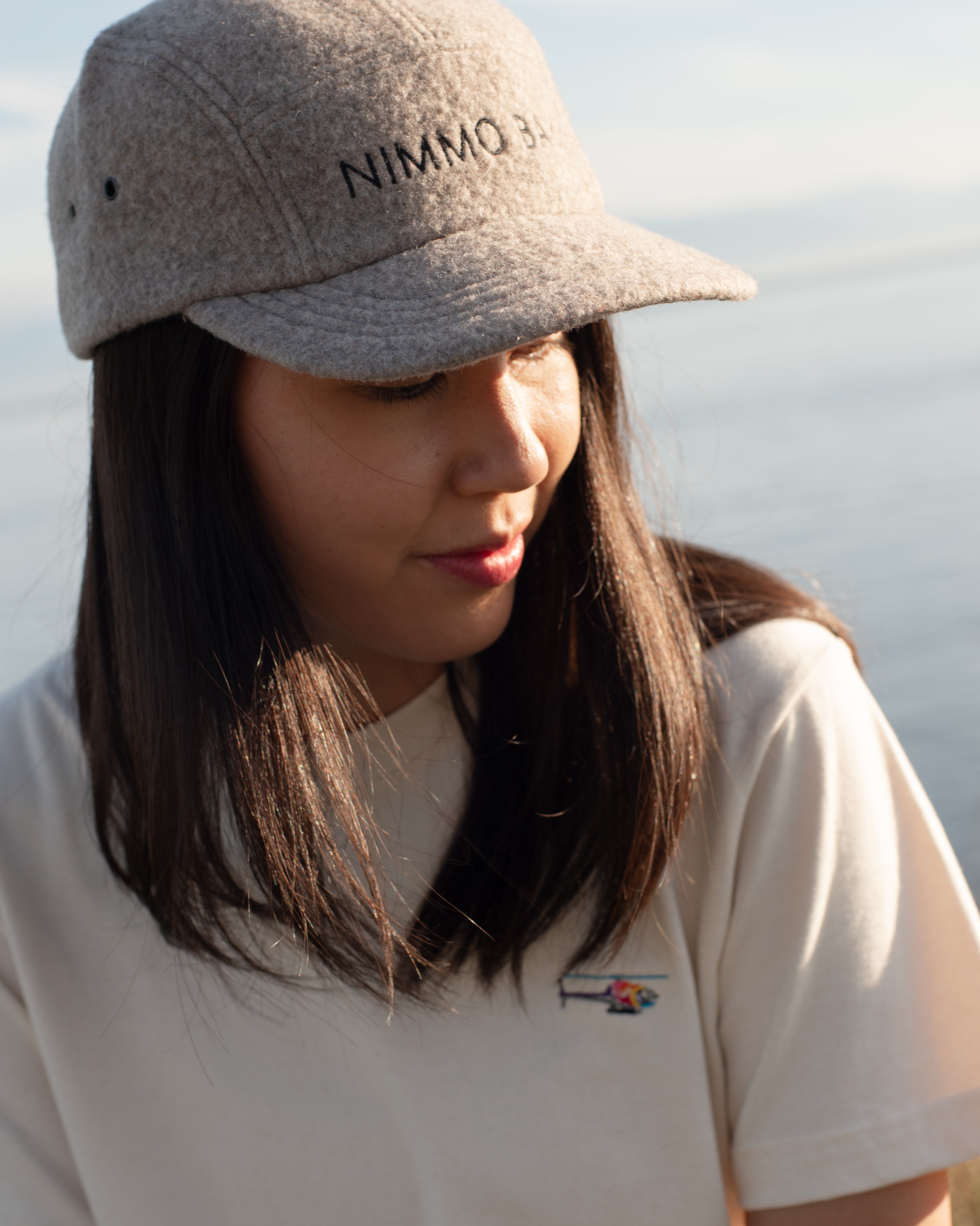A person wearing a beige anian fuzzy cap with the Nimmo Bay logo, an antique leather buckle, and an adjustable leather strap.
