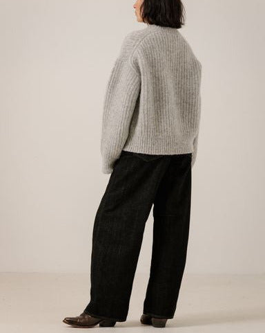 A model shown from the back wearing a light gray ribbed knit sweater and black wide-leg trousers against a plain off-white studio background.