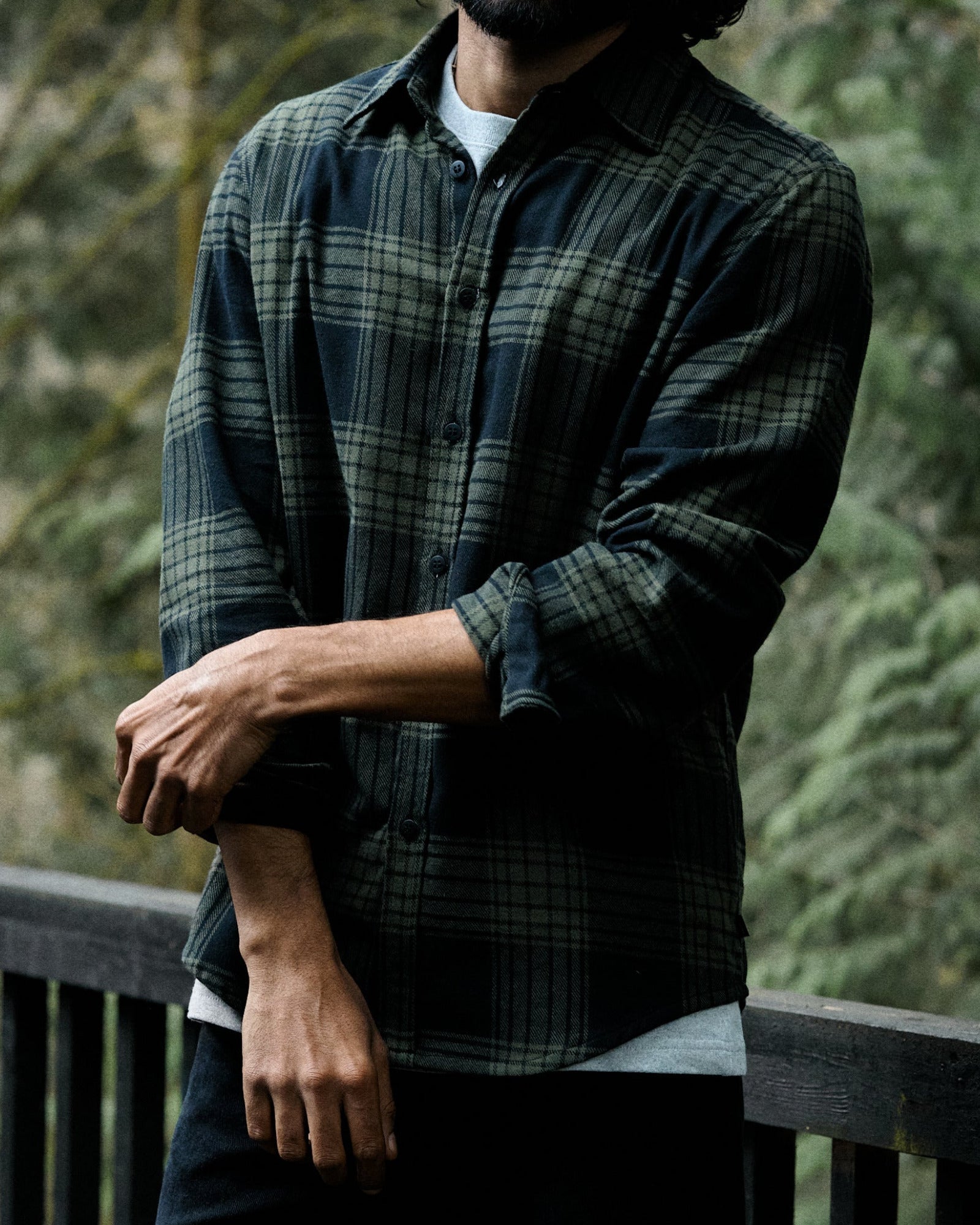 A man wearing a dark green and black plaid flannel shirt over a white undershirt, standing outdoors on a wooden deck with a blurred forest background.