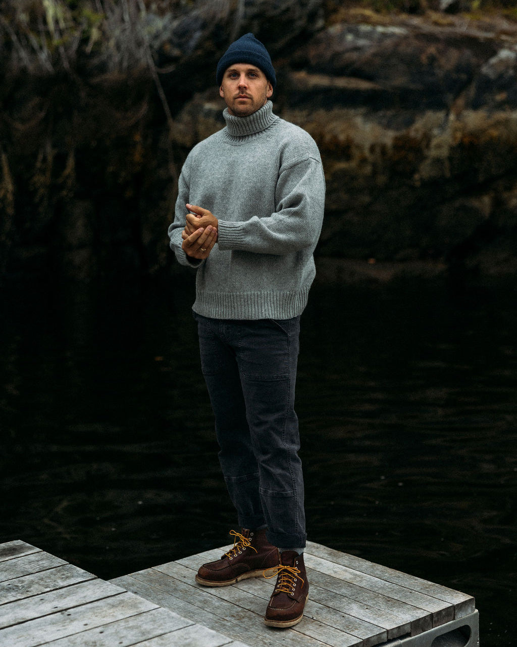 Man wearing a gray Bare Knitwear turtleneck sweater and dark pants standing on a wooden platform with a rocky background